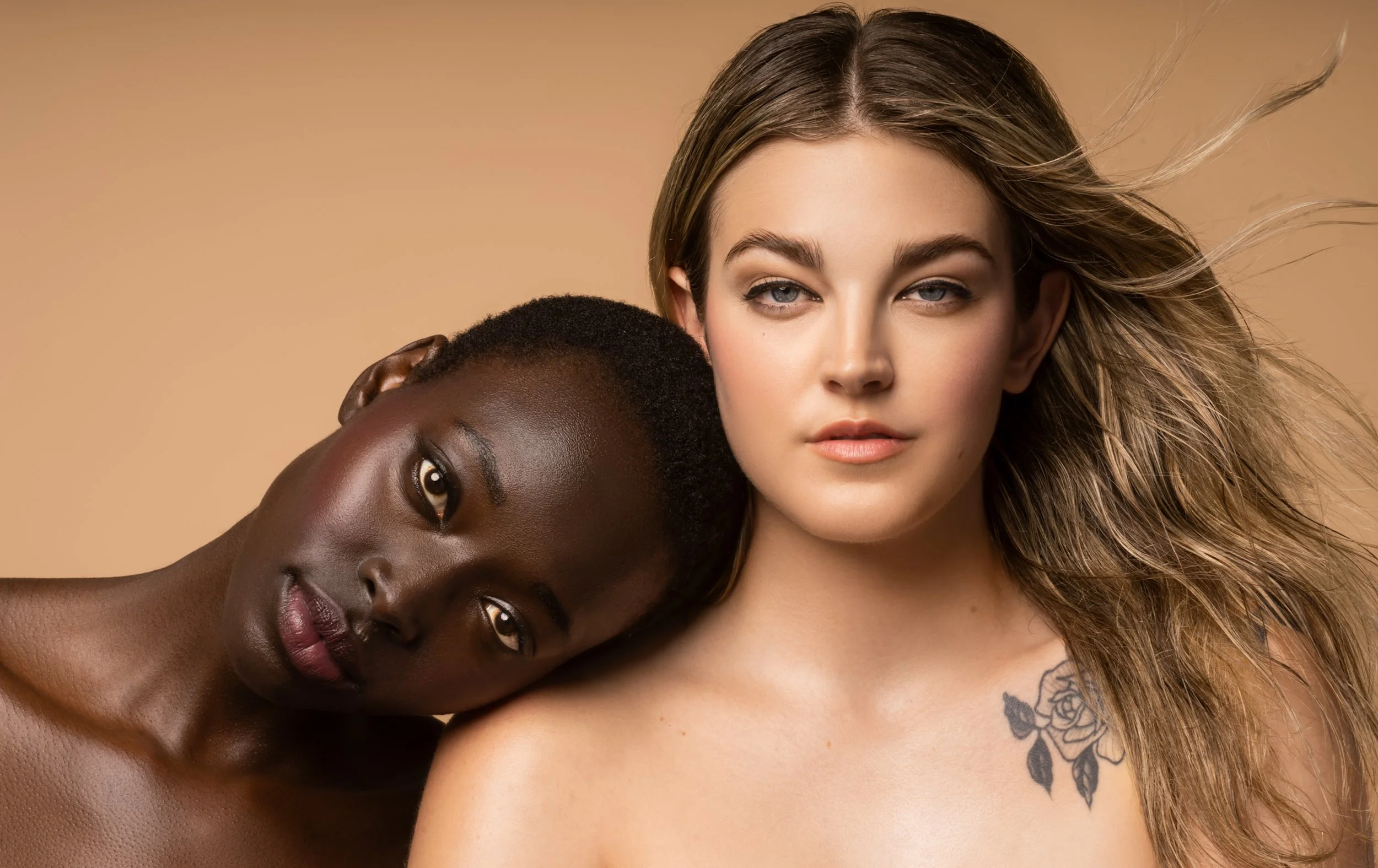 A woman with long, wavy blonde hair and light skin is seen with her head tilted slightly to the side, and a woman with short, dark skin and very short hair is resting her head on her shoulder, with her eyes closed. The background is a plain, light beige color.