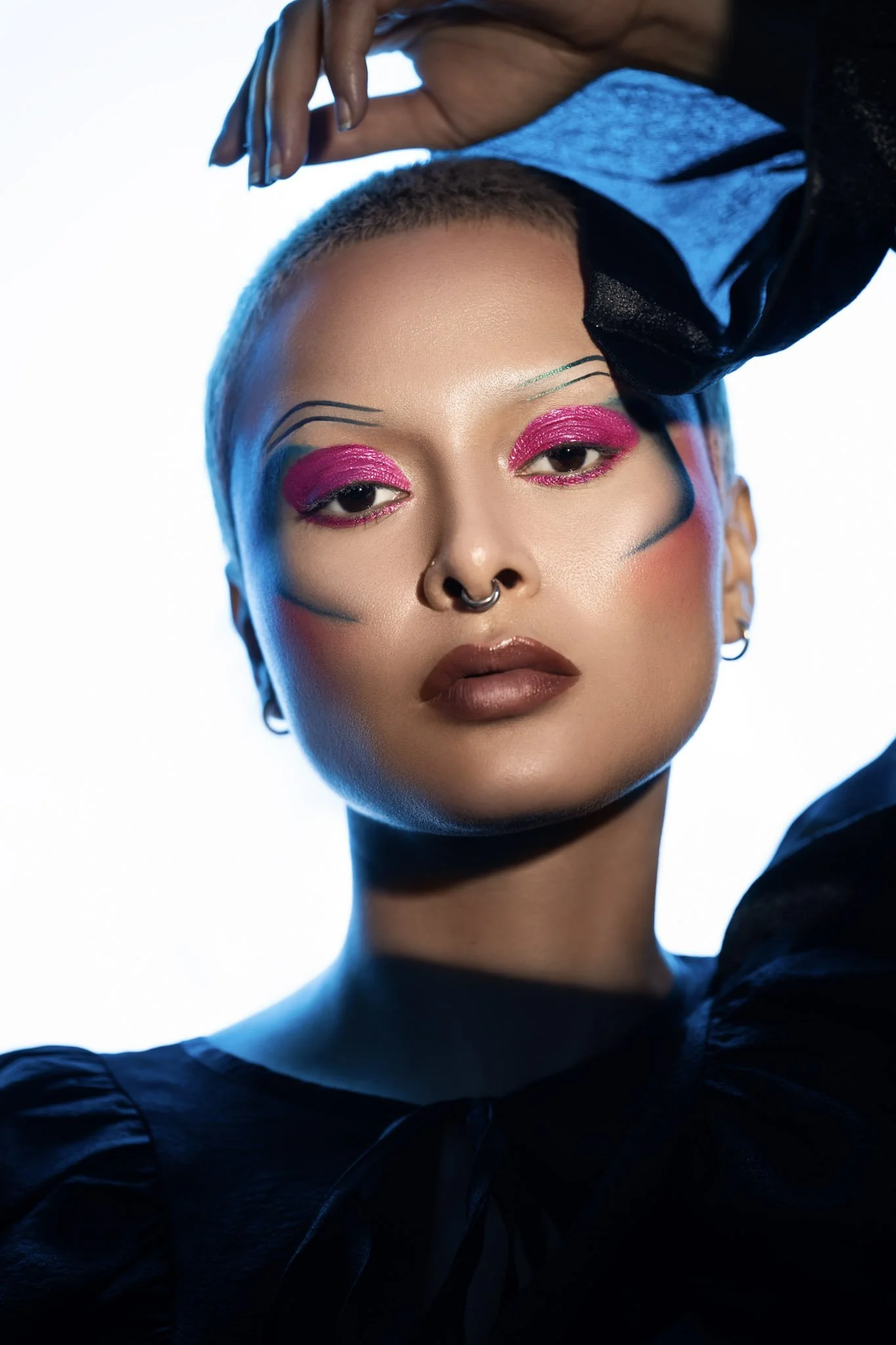 A woman with short hair and bold makeup, including bright pink eyeshadow and black graphic lines on her face, poses with her hand above her head against a white background. She wears a black top with puffed sleeves and has a septum piercing.