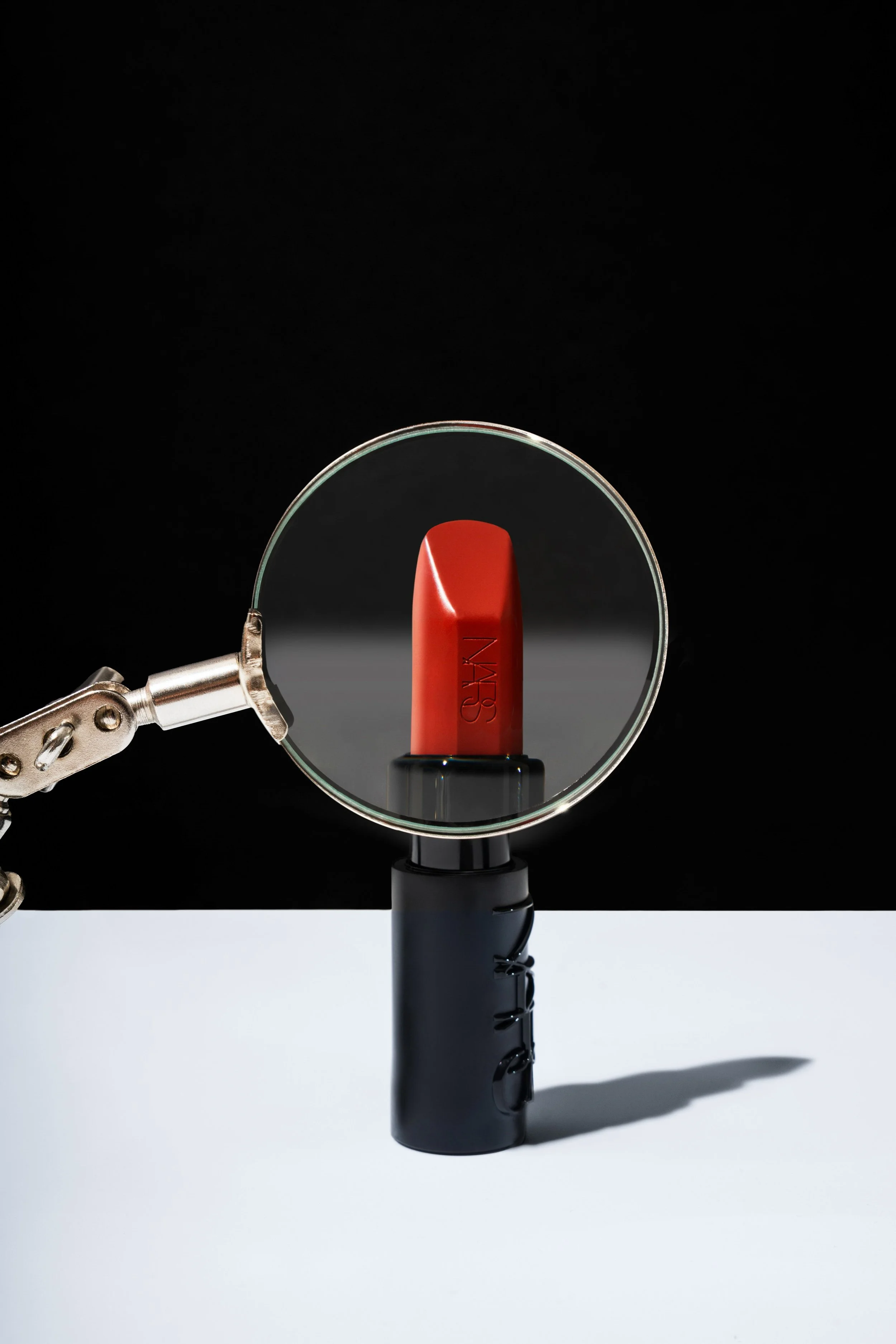 A black and white table with a red lipstick at center, a magnifying glass above it, and a black lamp at the base of the lipstick. The magnifying glass enlarges the lipstick and makes it appear prominent.