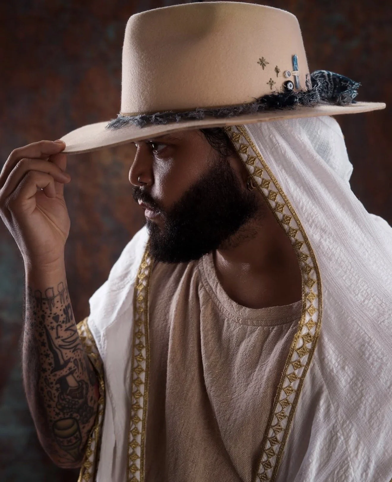 A man with a beard, tattoos on his arm, wearing a traditional white garment with gold embroidery, and holding a beige hat with pins and badges, tilting the hat to his face.