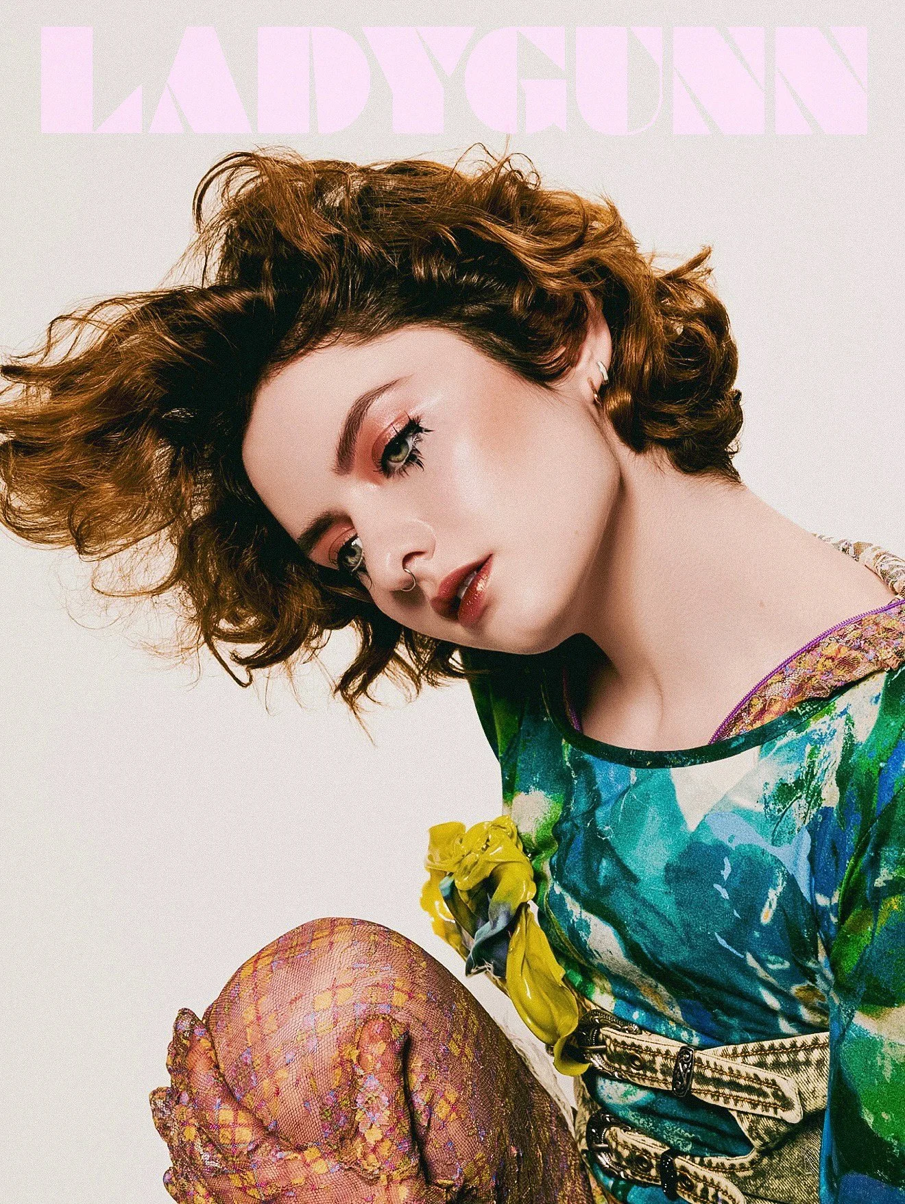 A young woman with curly brown hair and light makeup posing against a plain background. She wears a colorful, patterned dress with floral accents and fishnet stockings in bright, multicolored fabric. The image appears to be part of a magazine cover or advertisement.