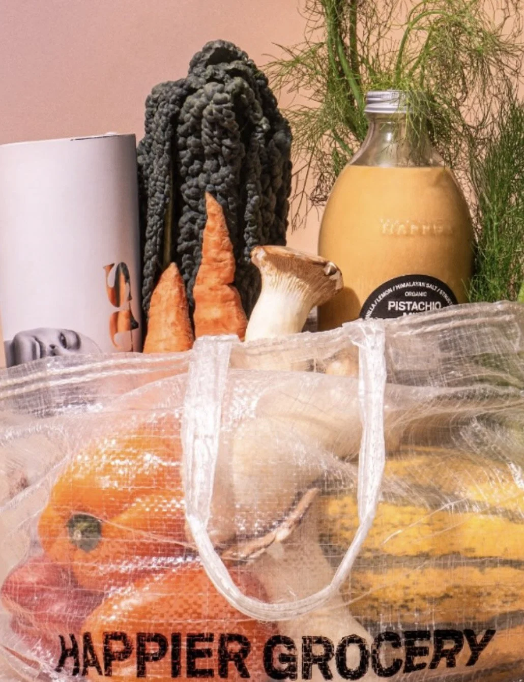 A reusable grocery bag with vegetables and a jar of pistachio nut spread on a table, with a decorative plant in the background.