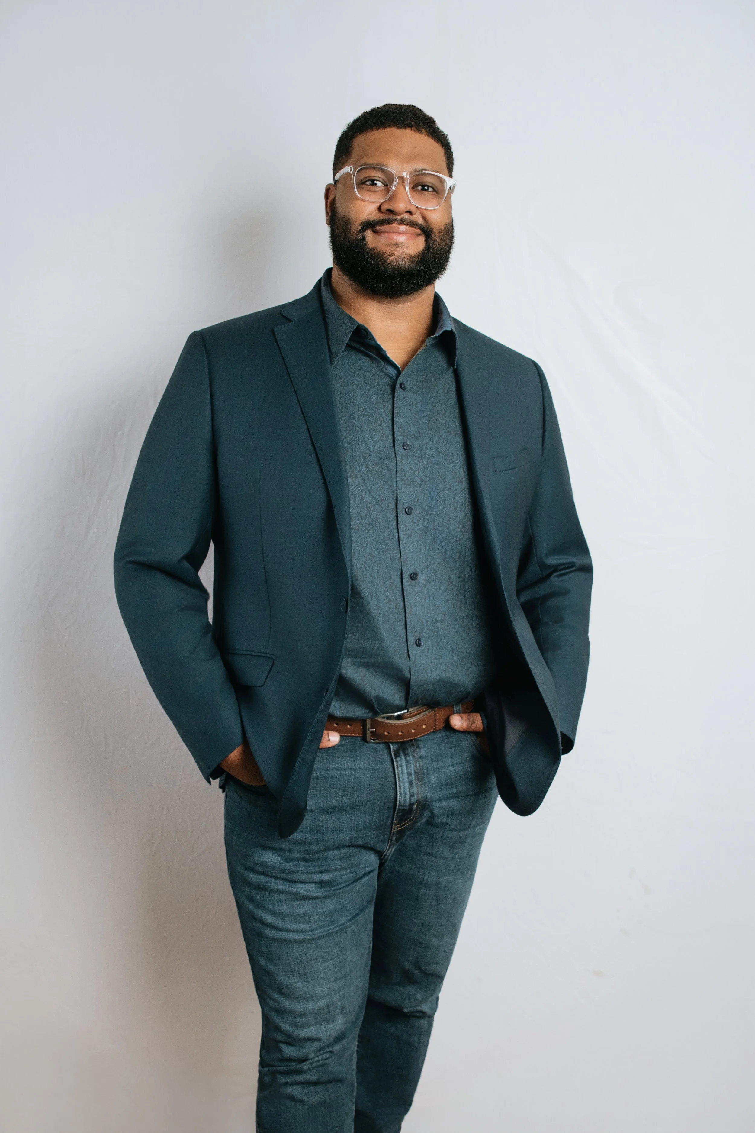 A man standing against a plain white wall, wearing glasses, a dark blazer, a patterned shirt, jeans, and a brown belt.