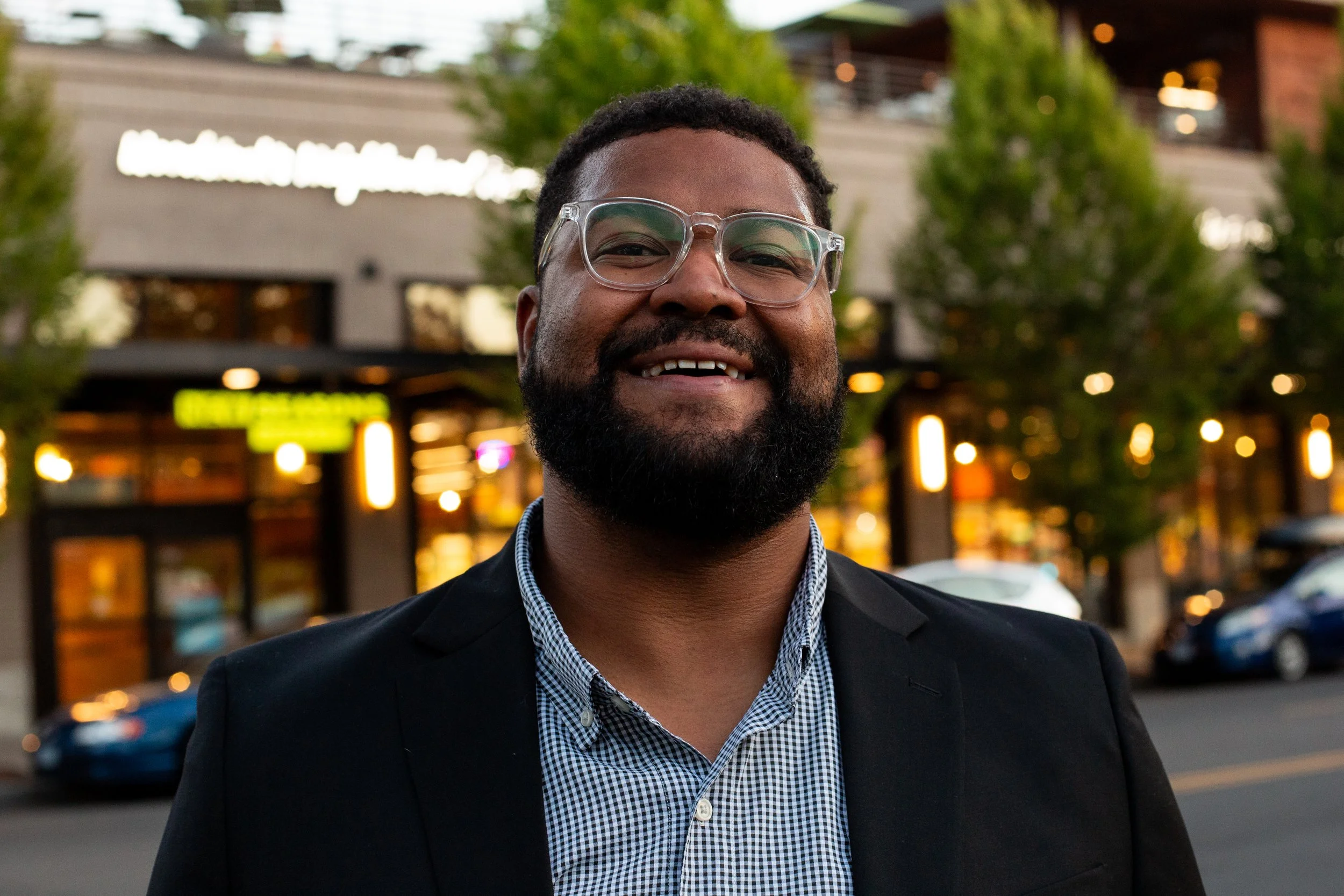 A man with a beard and glasses smiling outdoors, dressed in a black blazer and a checkered shirt, with a background of trees and a building with blurred lights.