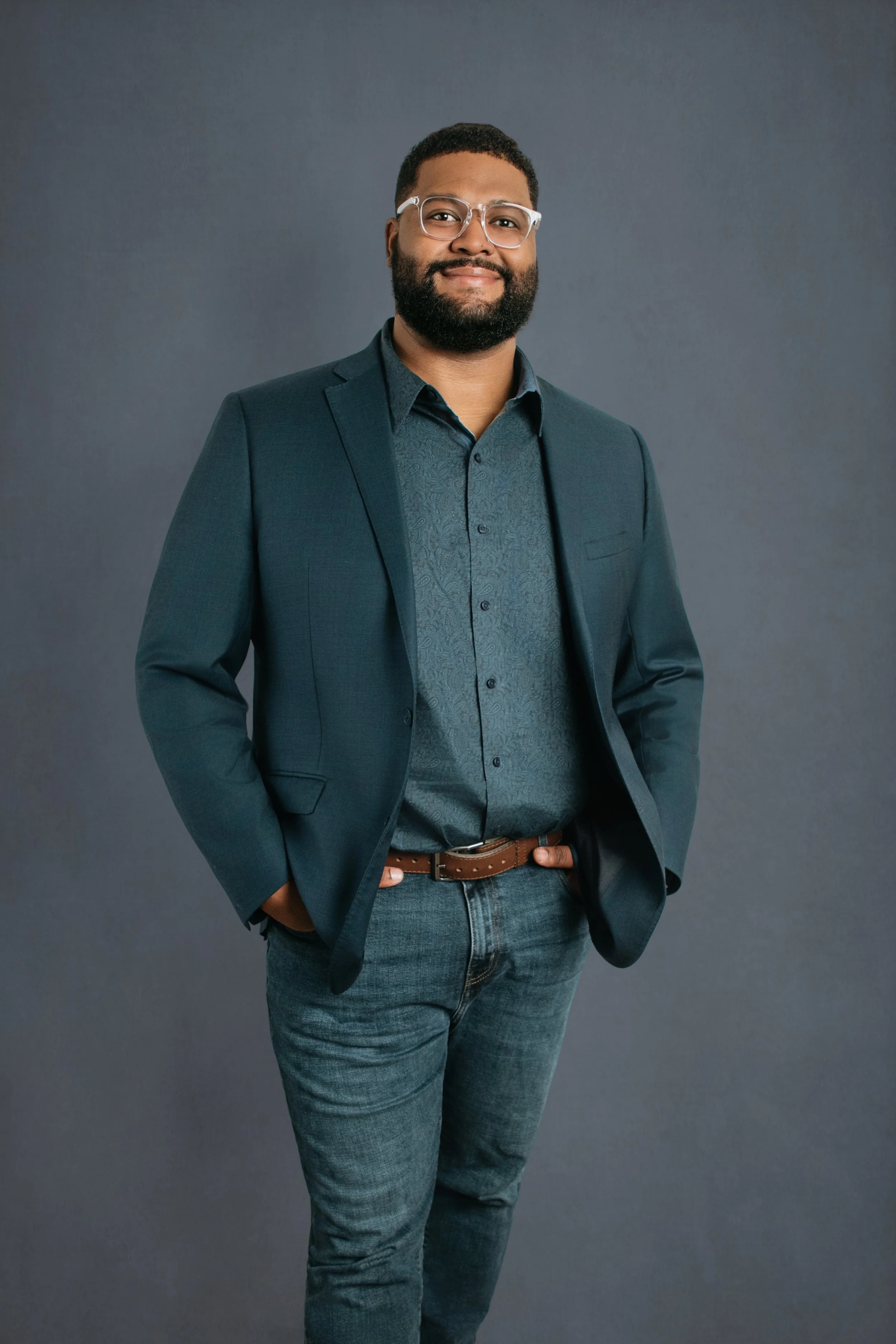 Portrait of a confidently smiling Black man with glasses, beard, dark blazer, patterned shirt, blue jeans, and a brown belt, standing against a plain gray background.