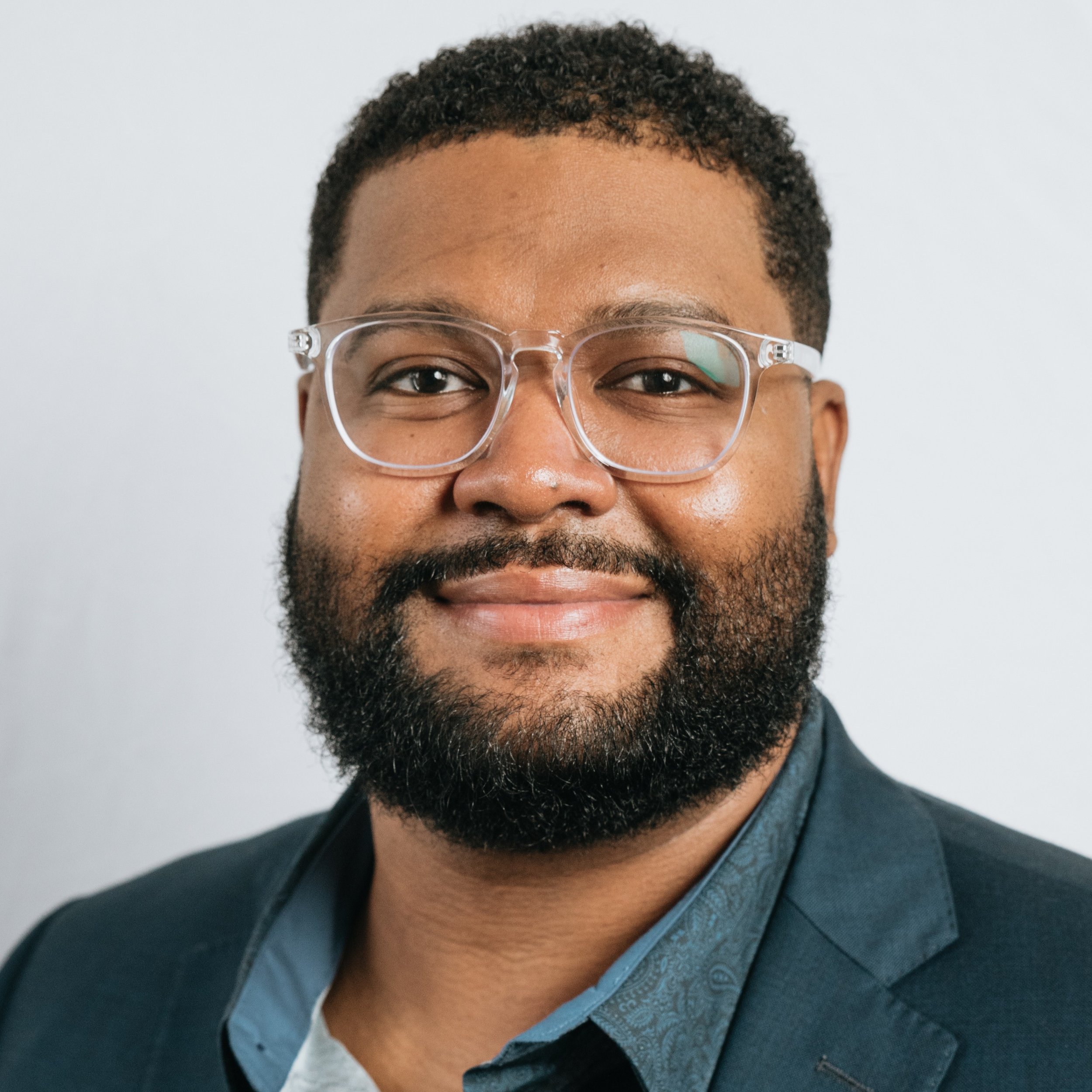 Close-up of a person's face showing the upper half, with curly short hair, glasses, a beard, and a smile, wearing a dark jacket and a blue shirt.