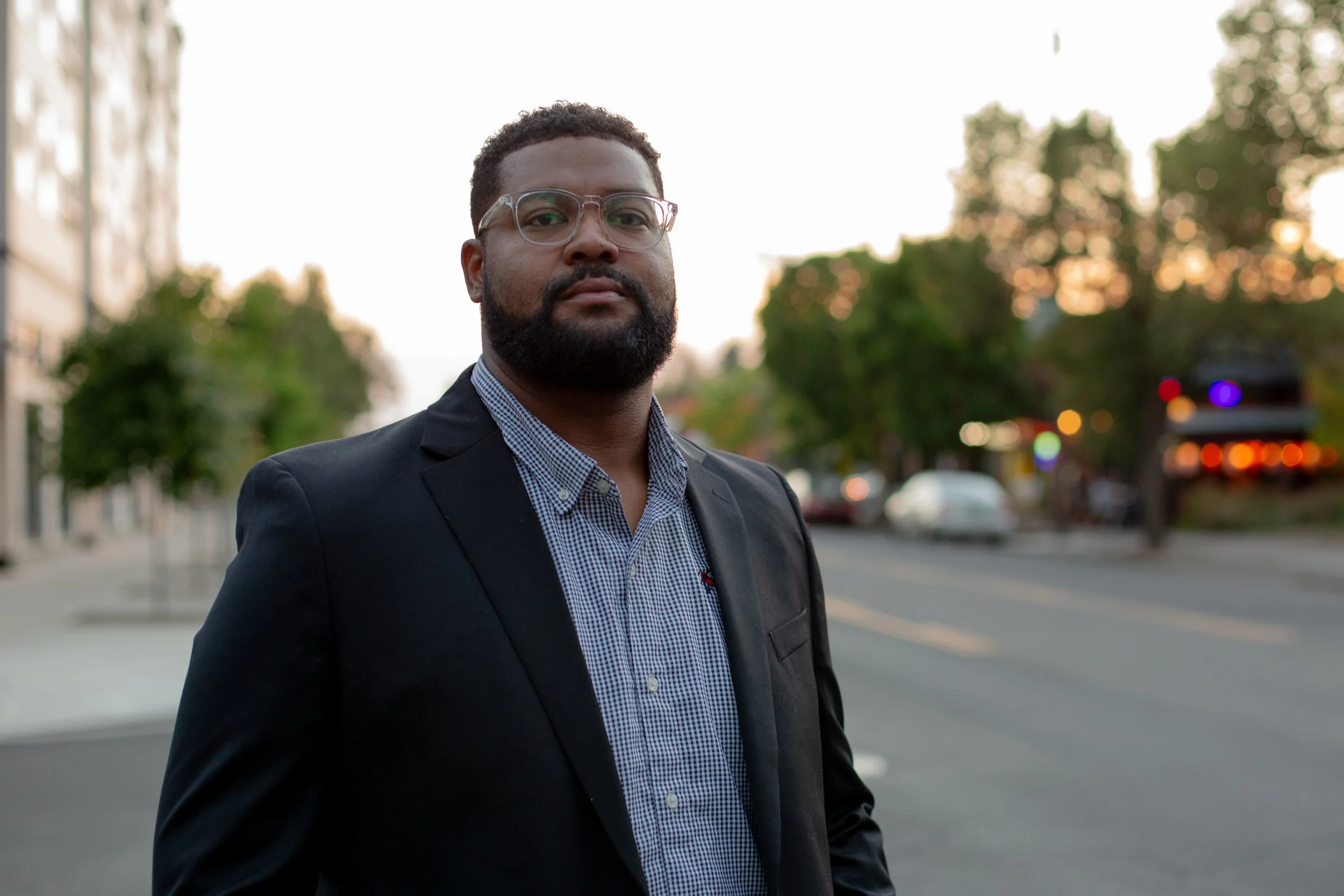 A Black man with a beard and glasses in a business suit standing on a city street at dusk, with trees and blurred city lights in the background.