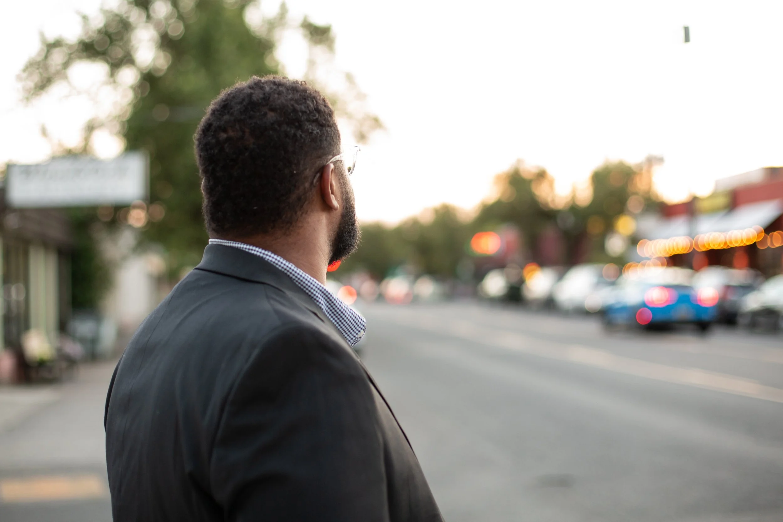 A man with short, curly hair and a beard, wearing glasses and a dark suit with a checkered shirt, is standing on a street looking towards the bright background of the evening sky, surrounded by blurred lights and trees.