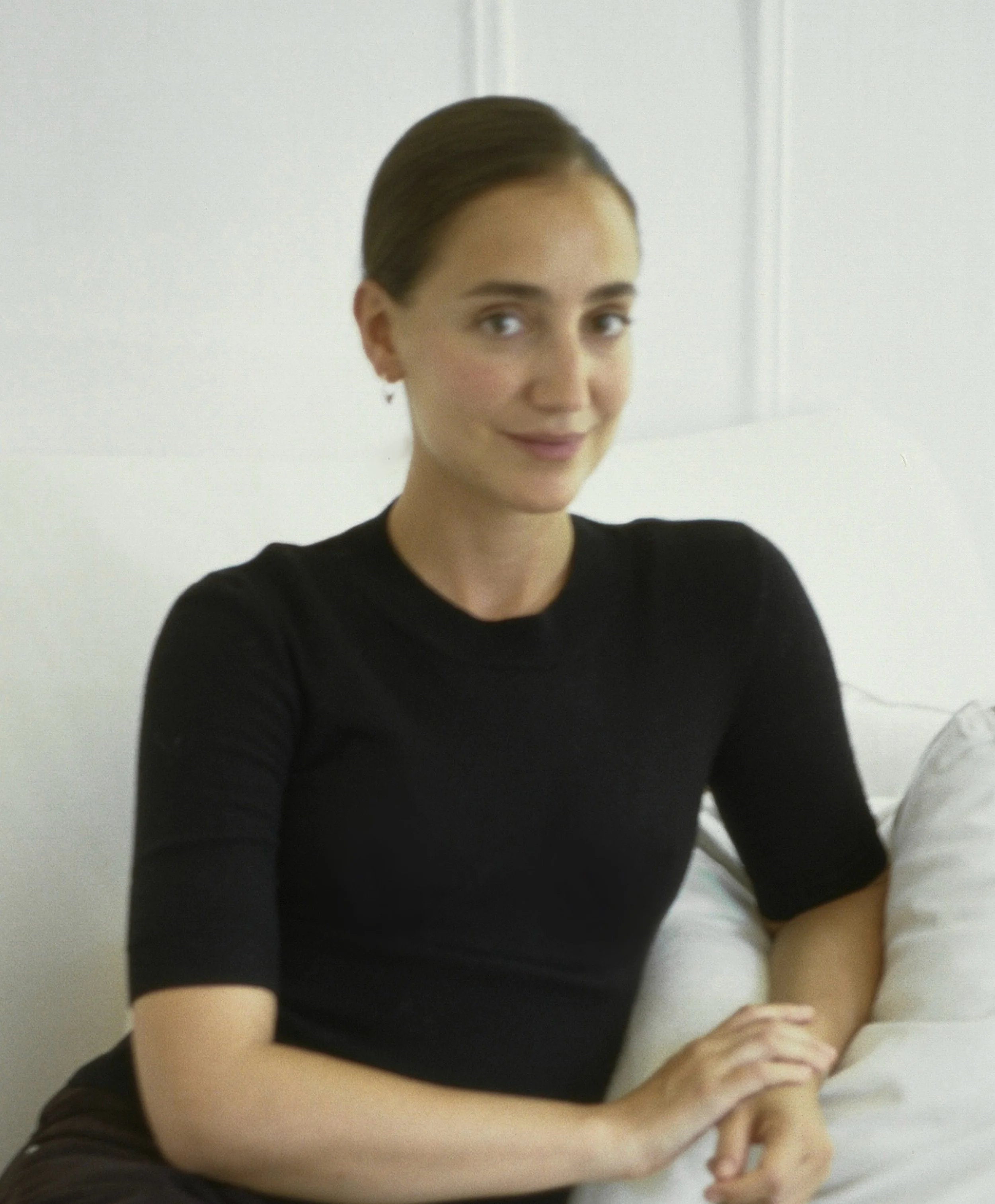 A woman with her hair swept back, wearing a black shirt, sitting at a white table and looking at the camera with a slight smile.