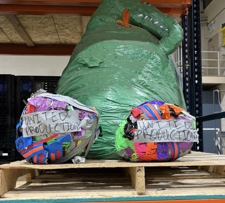Large green inflatable or sculpture resembling a dinosaur or dragon with orange nose and long tail, placed on a wooden pallet. Two smaller colorful items with handwritten signs reading 'United Productions' are in front of it.
