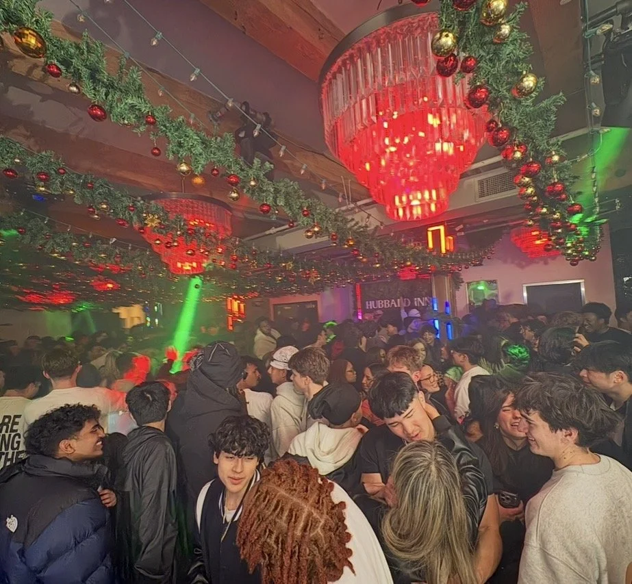 Crowd of young people dancing and socializing in a party space decorated with Christmas ornaments and garlands, with large red chandelier lights and green and red lighting.