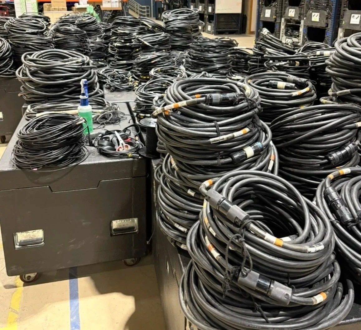 Stacks of coiled black cables on tables in a workshop or storage room, with some electronic equipment and tools visible in the background.