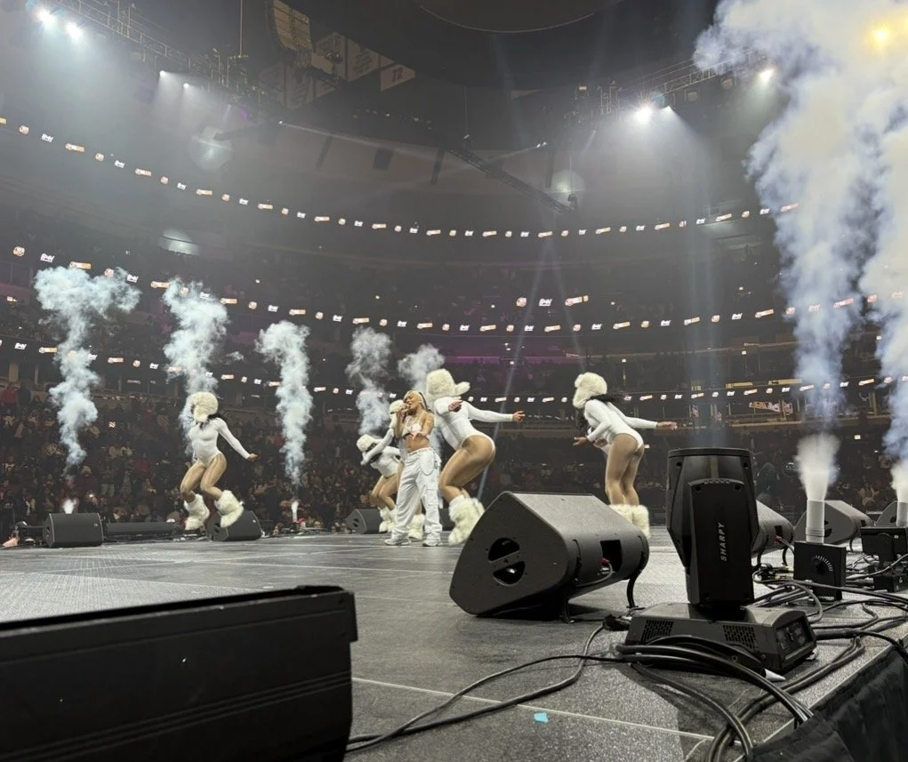 Performers on stage dressed in white winter-themed costumes with puffball hats, surrounded by clouds of smoke and bright stage lights, in an indoor concert arena.