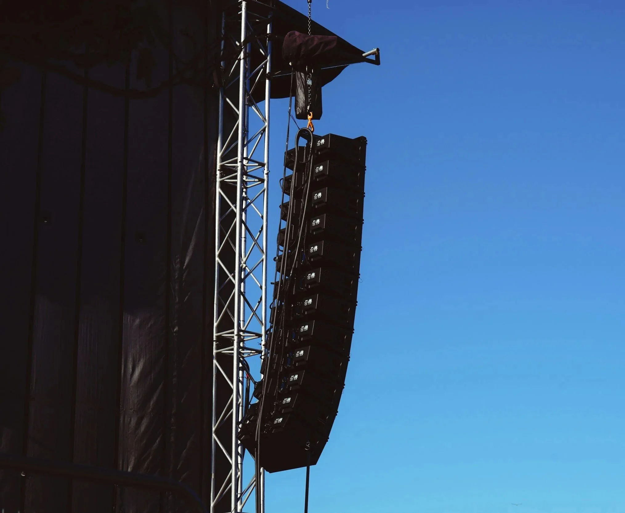 A large outdoor speaker system is mounted on a metal scaffold against a blue sky, likely at a concert or event.