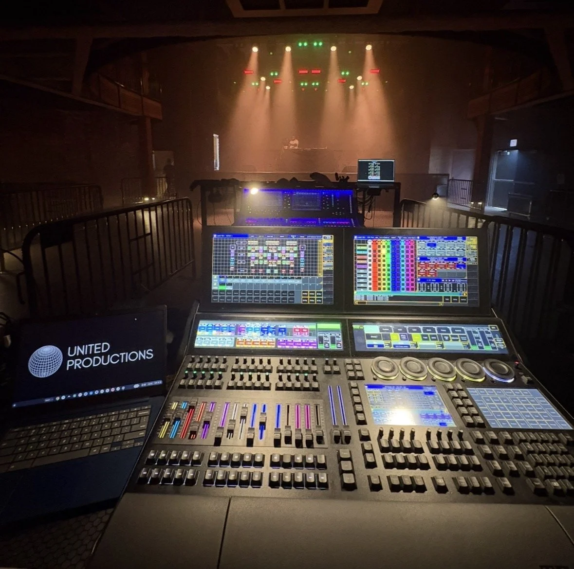 A sound mixing board and computer screens with colorful audio interface displays in a dark concert venue or club, with a stage lit with green and red lights in the background.