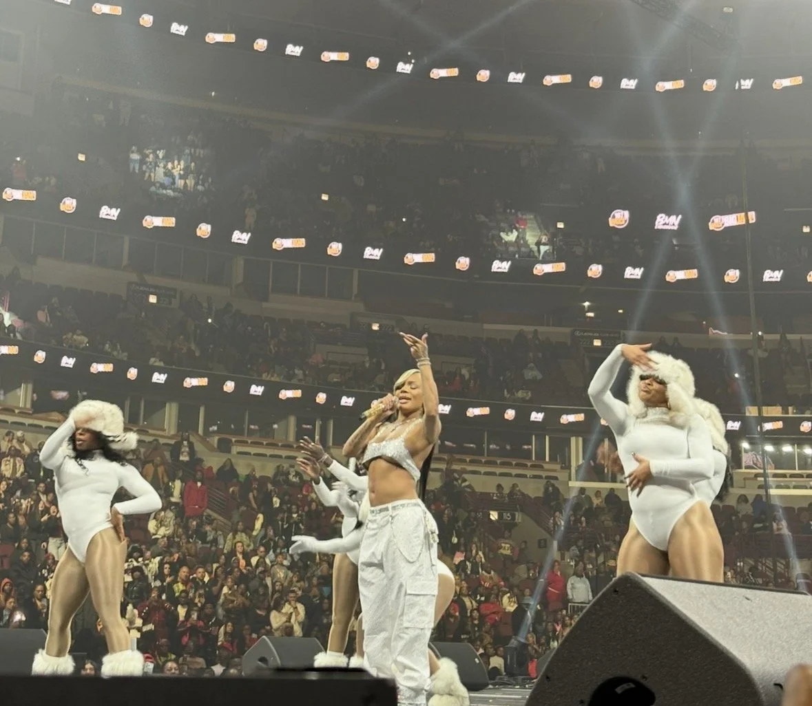 A live stage performance with a female singer and backup dancers dressed in winter-themed costumes in a large arena filled with audience.