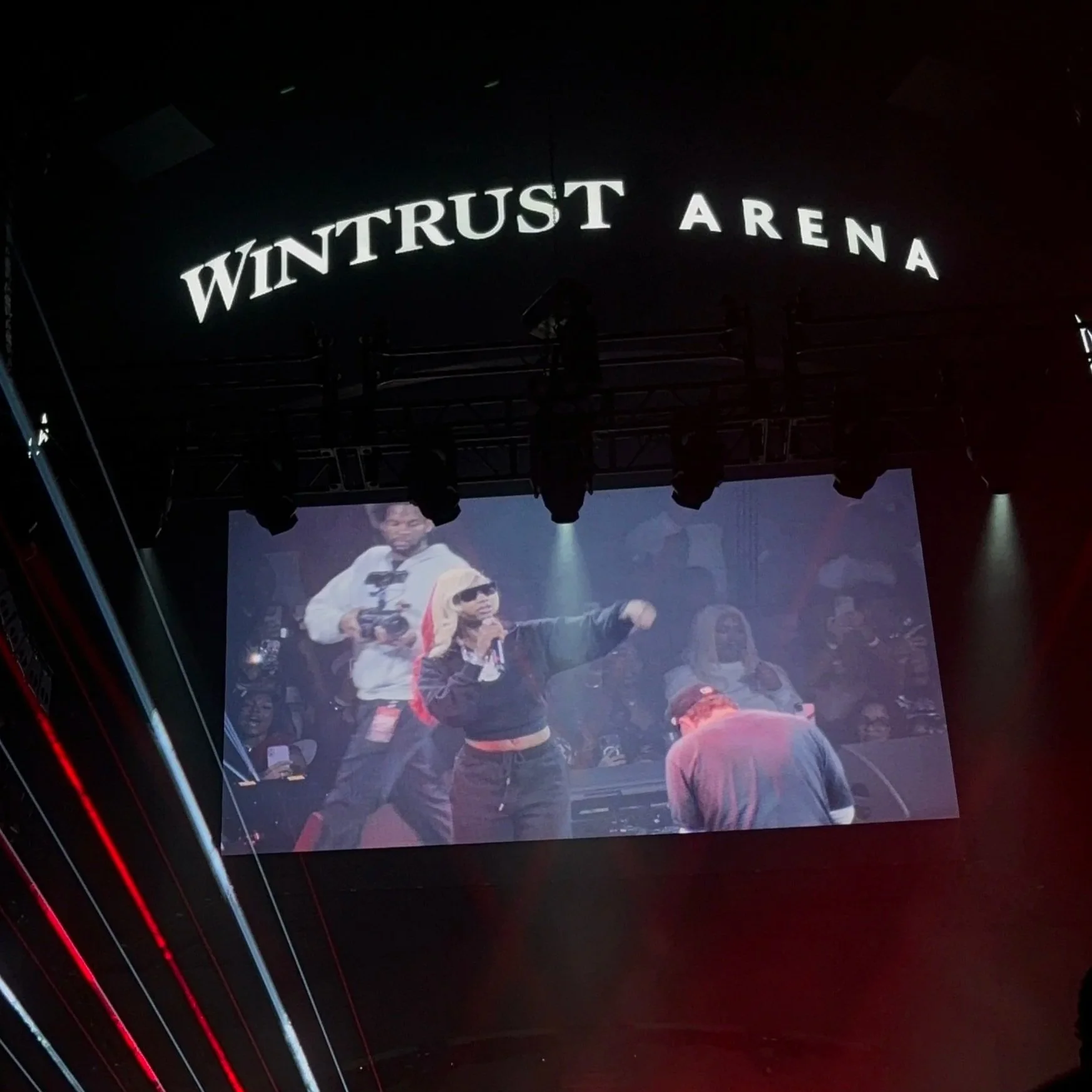 Large indoor auditorium with a stage displaying a projection of two young men wearing hoodies, one of whom is holding a microphone. The backdrop shows the name 'Wintertrust Arena' in illuminated letters.