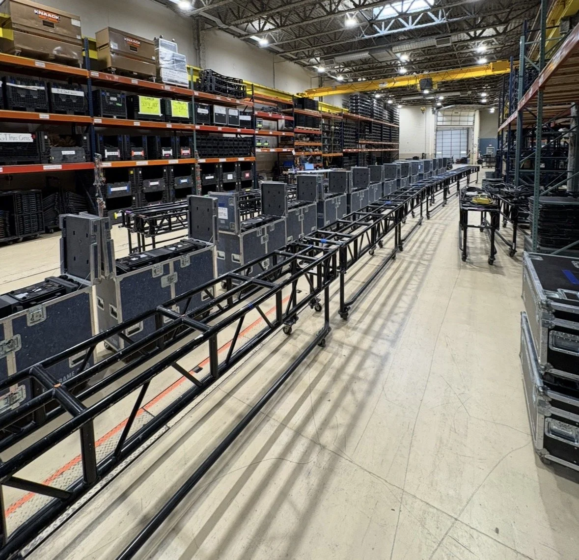 Interior of a warehouse with shelves stacked with electronic equipment and black storage cases, and a long table or cart with electronic devices in the center.