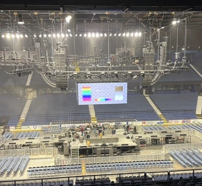 Empty indoor arena or stadium with a stage setup for an event, large screen displaying test pattern, and rows of seats surrounding the stage, with equipment and lighting rigs overhead.