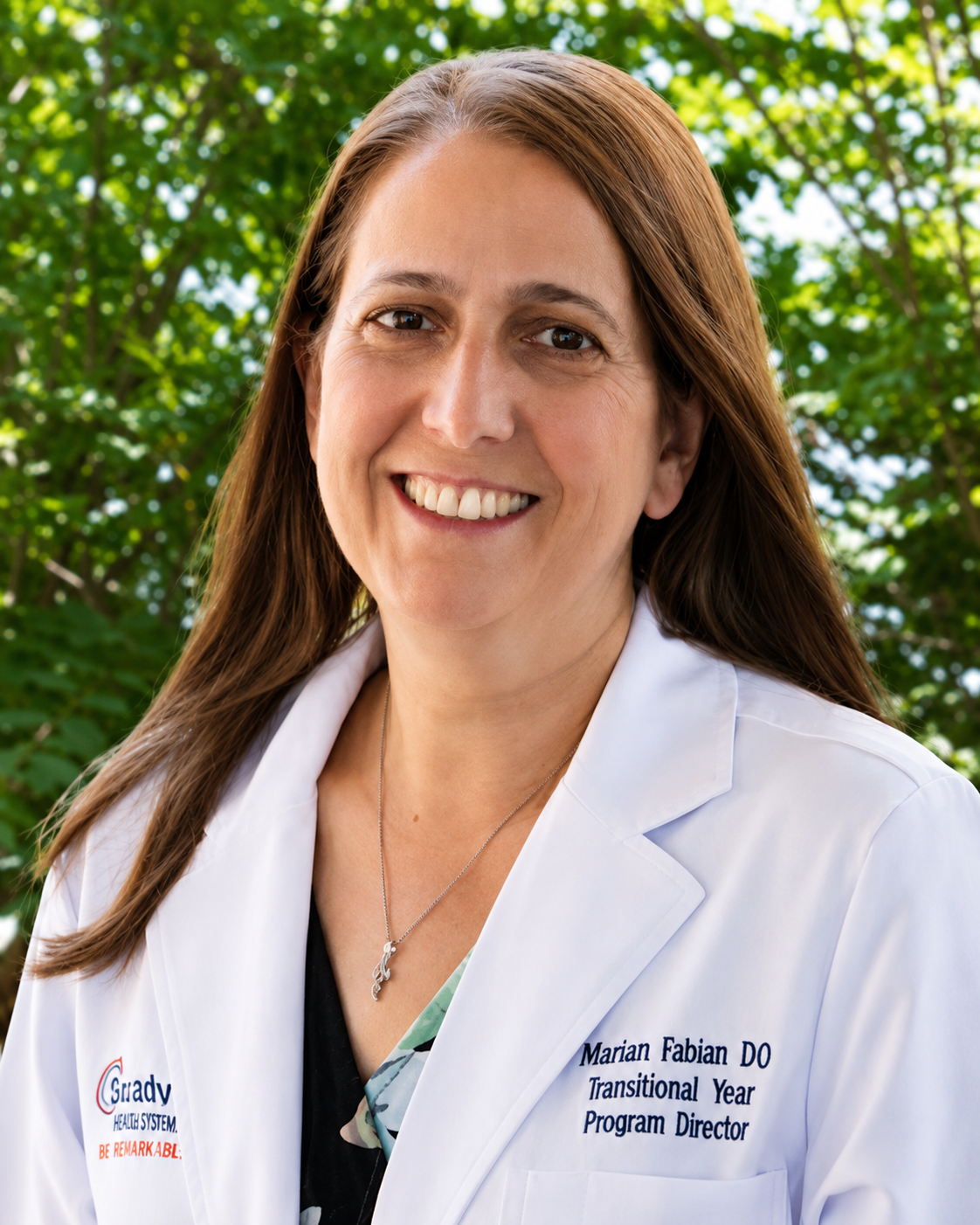 Portrait of a woman with brown hair, wearing a white lab coat with her name and title and smiling in an outdoor setting with green foliage in the background.