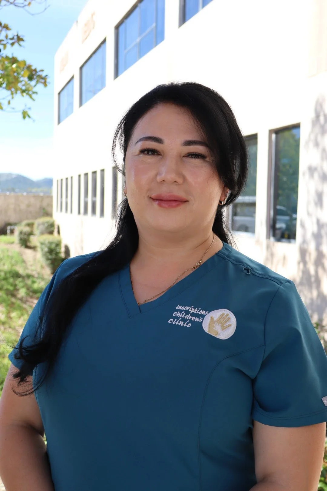 Woman in blue scrubs standing outdoors in front of a modern white building with large windows, sunny sky, and trees in the background.