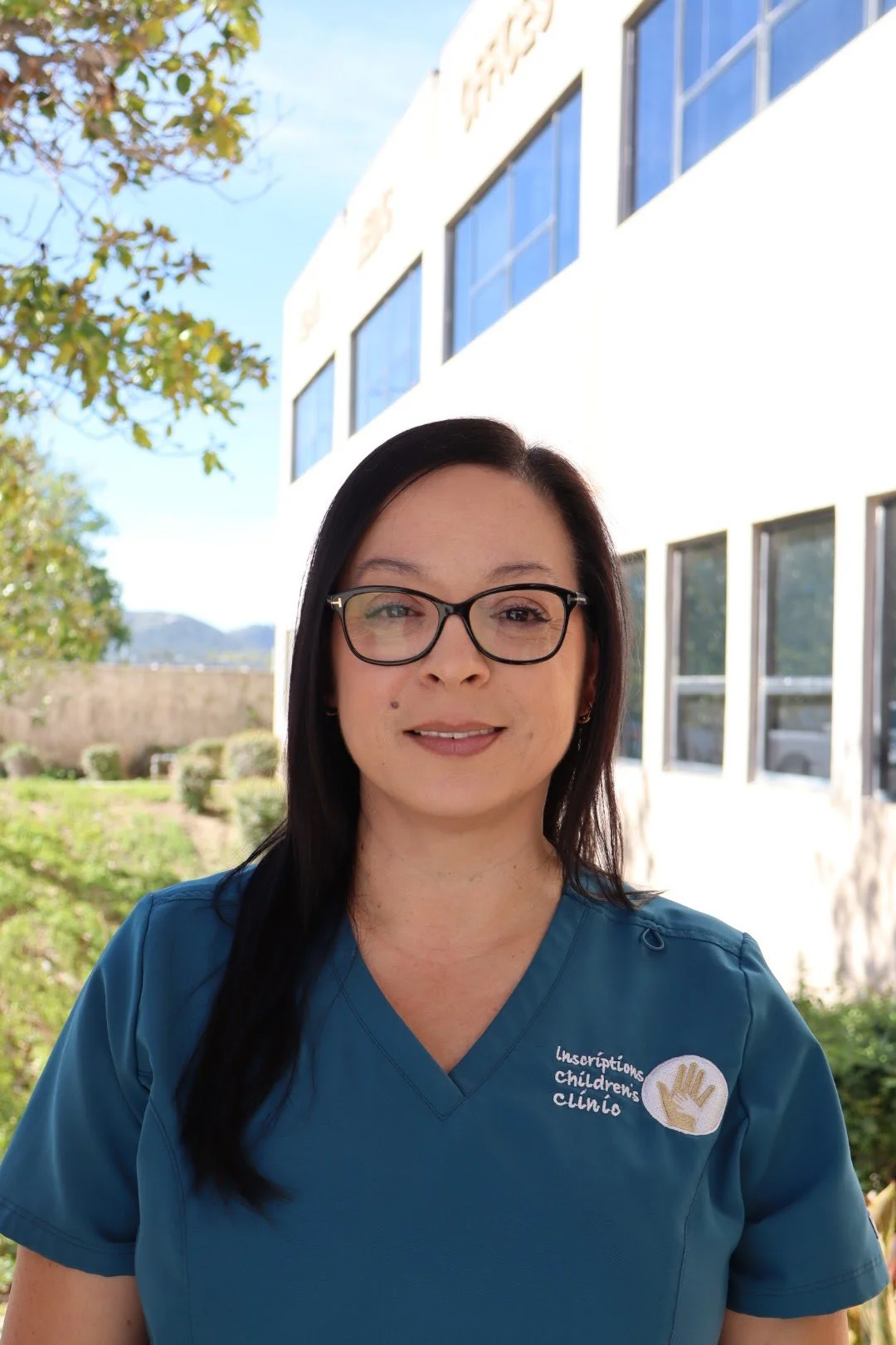 A woman in medical scrubs standing outside in front of a white building with large windows, under a tree with green leaves, sunny day with clear sky.