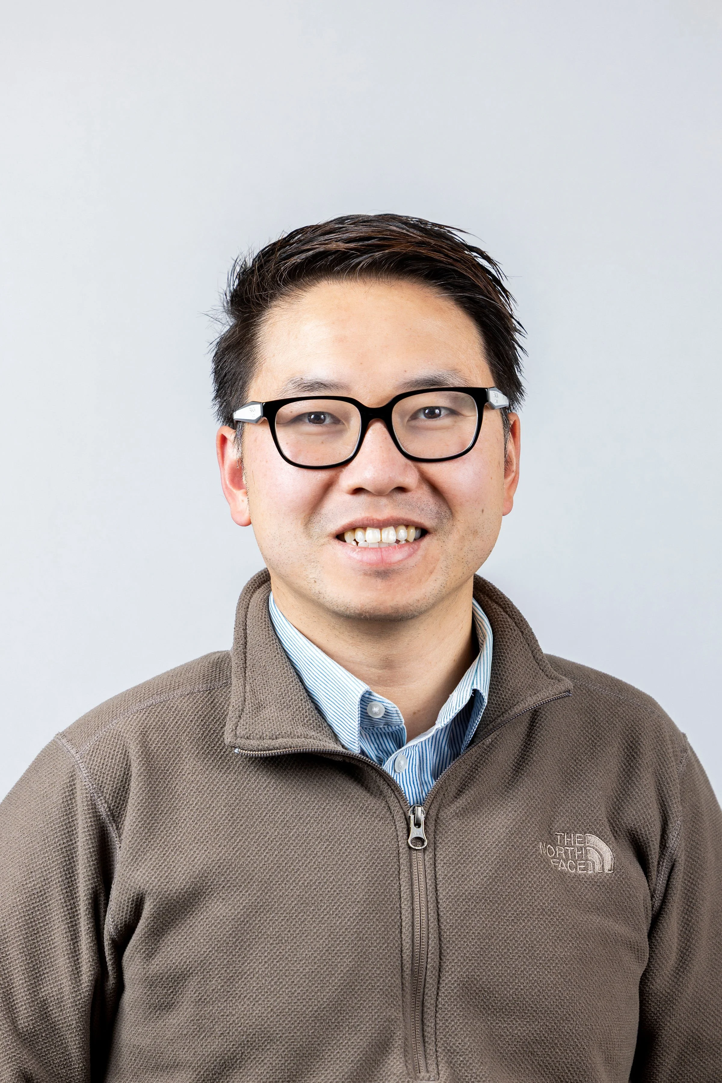 A smiling man wearing glasses, a collared shirt, and a brown North Face jacket against a plain light background.