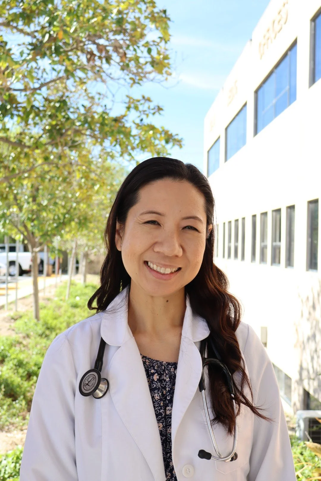 A female doctor or healthcare professional outdoors with a white coat and stethoscope, smiling at the camera, with trees and a building in the background.