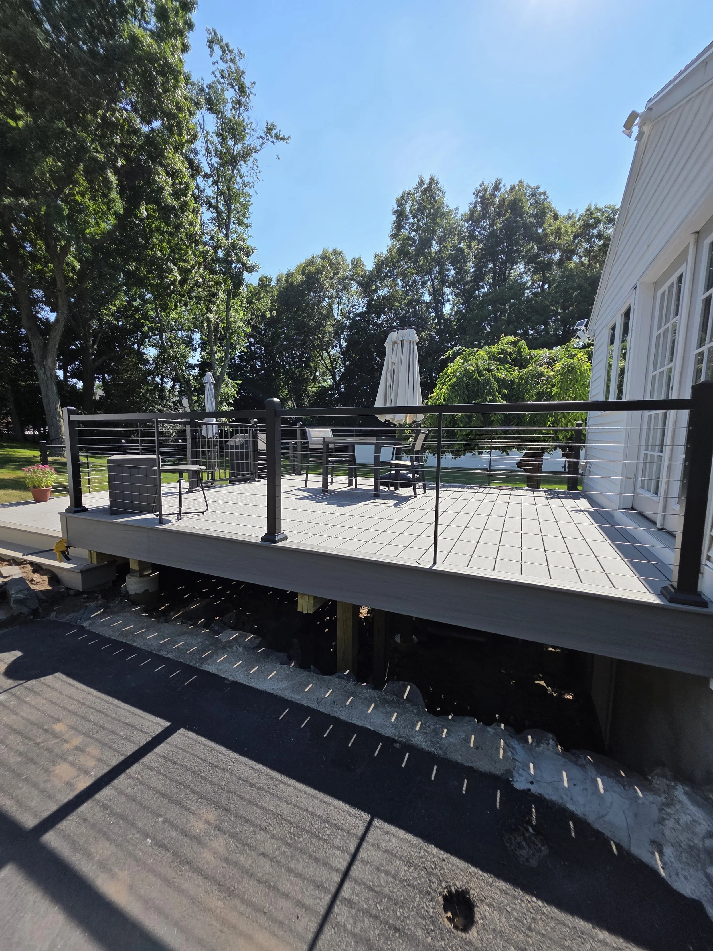 A wooden deck with black railings and patio furniture, surrounded by green trees and a white house, under a clear blue sky.