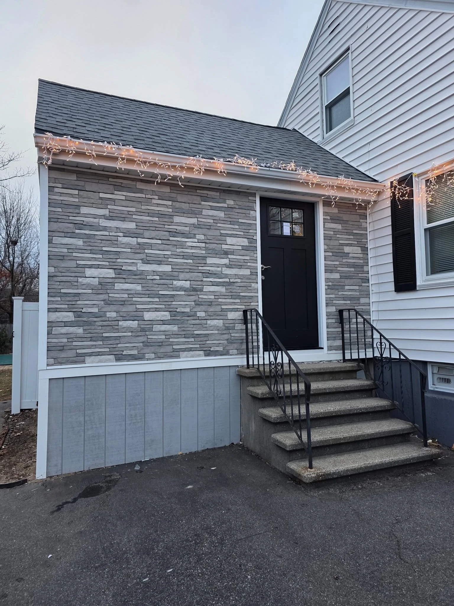 Front of a house with a black door, stone veneer siding on the lower half, white shingle siding on the upper half, and string lights hanging along the roofline. Concrete stairs with a black metal railing lead to the entrance, and the ground in front is paved with asphalt.