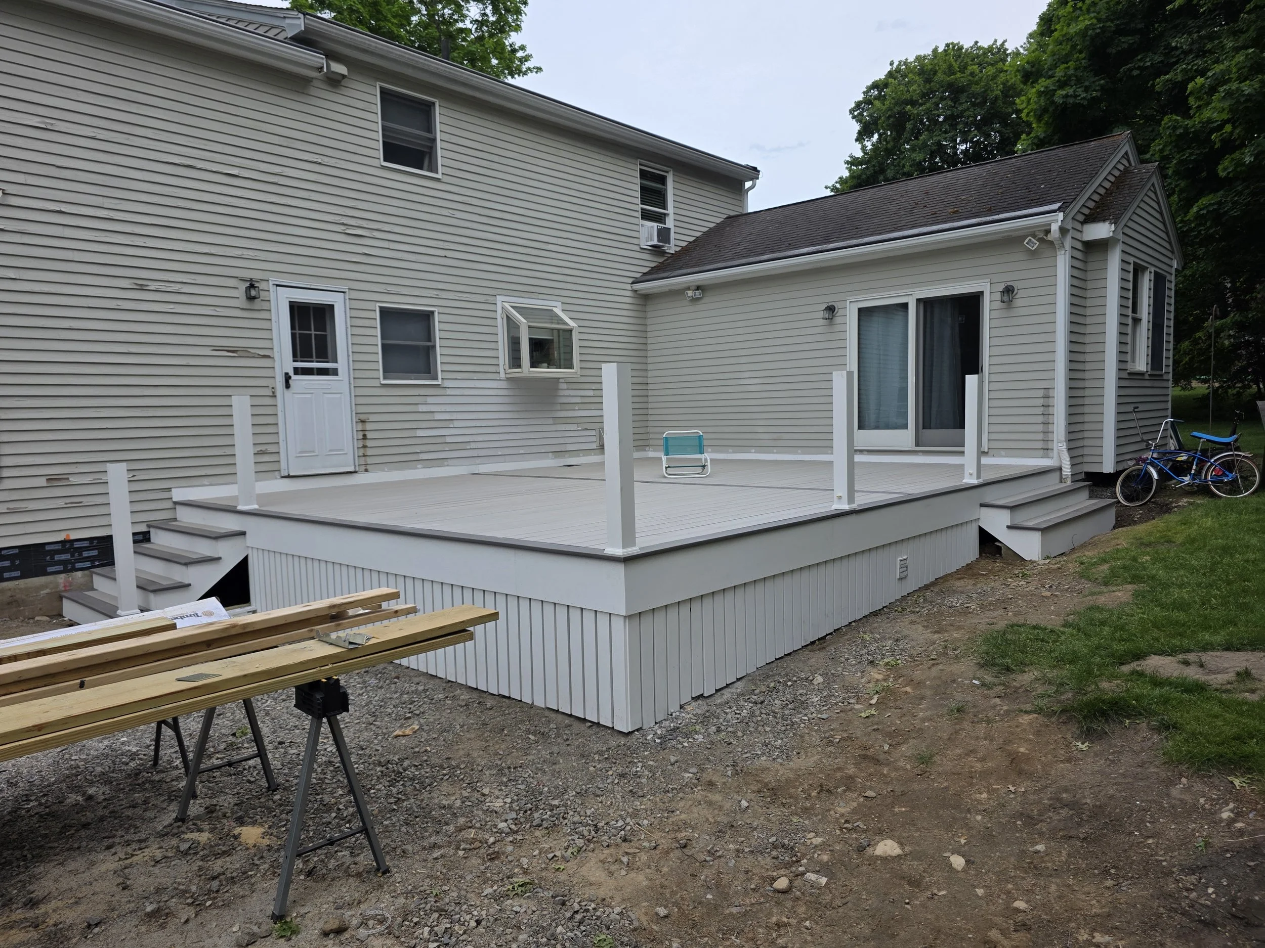 Newly constructed backyard deck with stairs, adjacent to a house with beige siding and multiple windows, surrounded by green trees.
