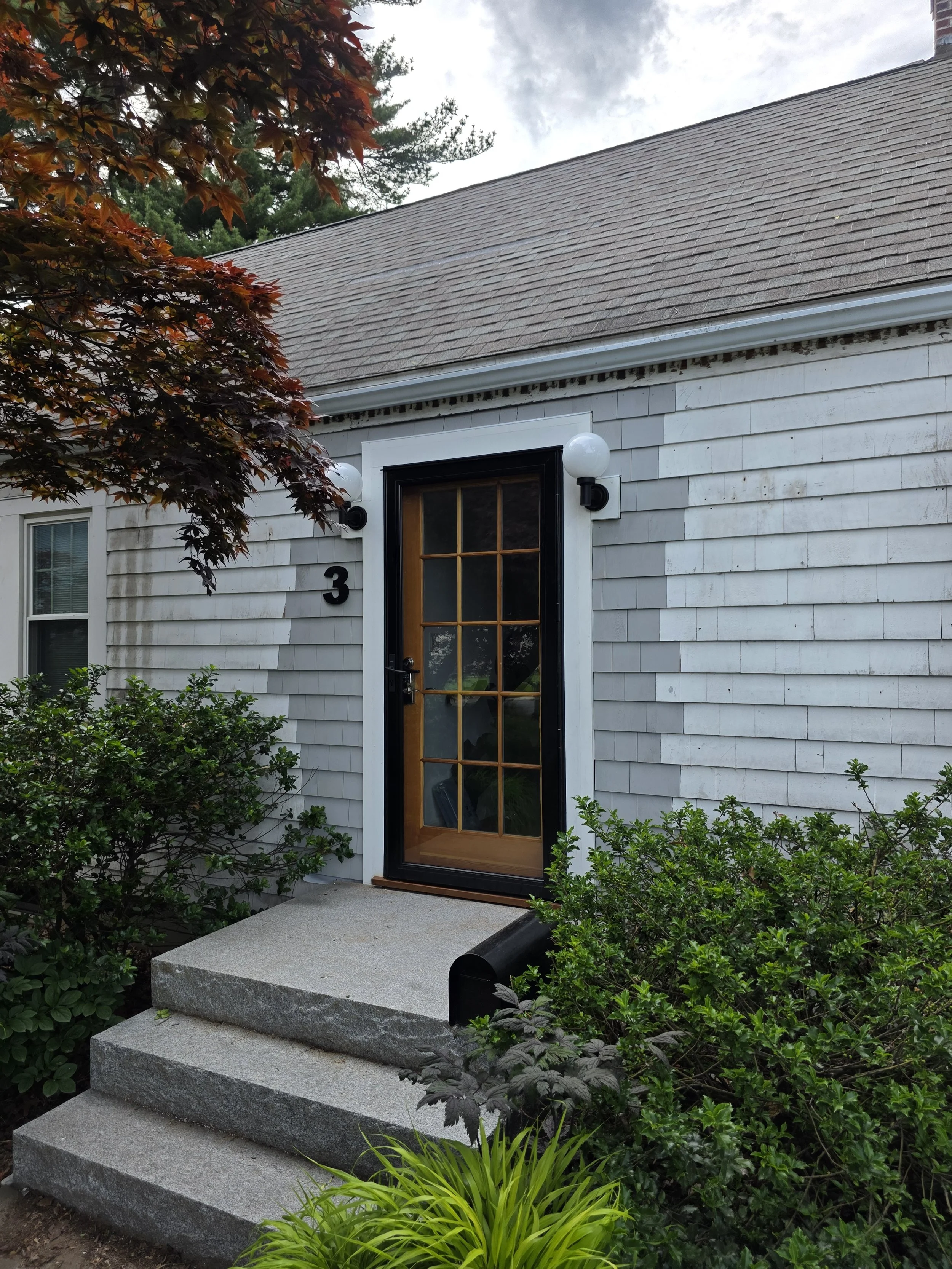Front entrance of a house with a wooden door, white siding, and two exterior lights. There are four concrete steps leading to the door, surrounded by green bushes and plants. The house has a gray roof and there is a tree with reddish leaves on the left side of the image.