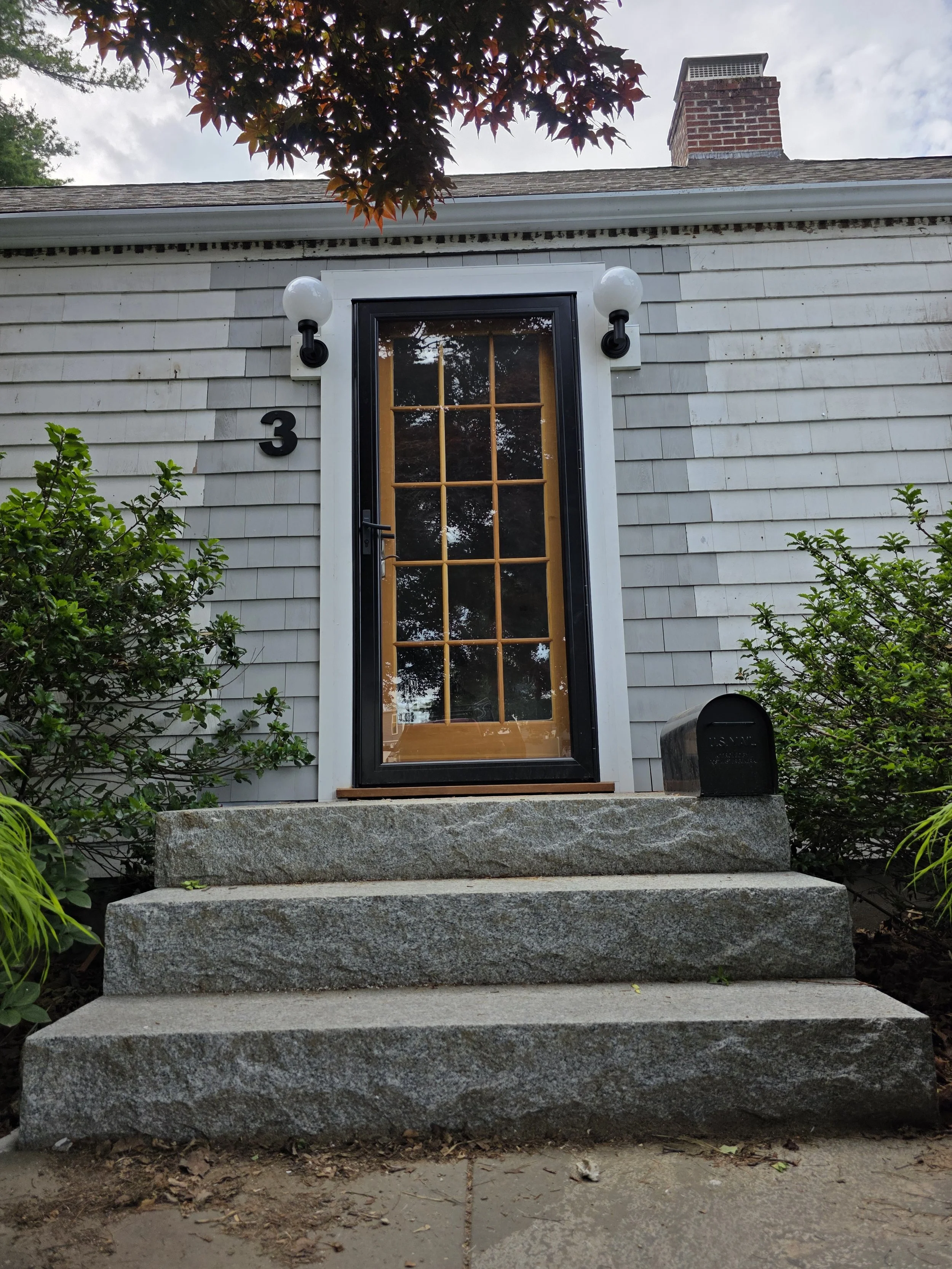 Front porch of a house with gray shingle siding and steps made of large gray stones. The house has a black door with glass windows, and two black outdoor wall lights on either side of the door. The house number '3' is displayed to the left of the door. Some green bushes are on either side of the steps, and a mailbox is visible on the right side. The roof has a brick chimney, and a tree with reddish-brown leaves partially shades the scene. The sky is cloudy.