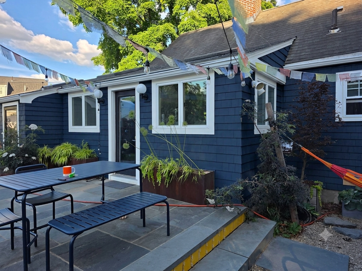 A backyard patio with a black metal table and matching benches, surrounded by potted plants. Colorful paper lanterns and bunting decorate the area, with a house painted blue with white trim in the background. A tree and a hammock are visible, with clear blue skies overhead.