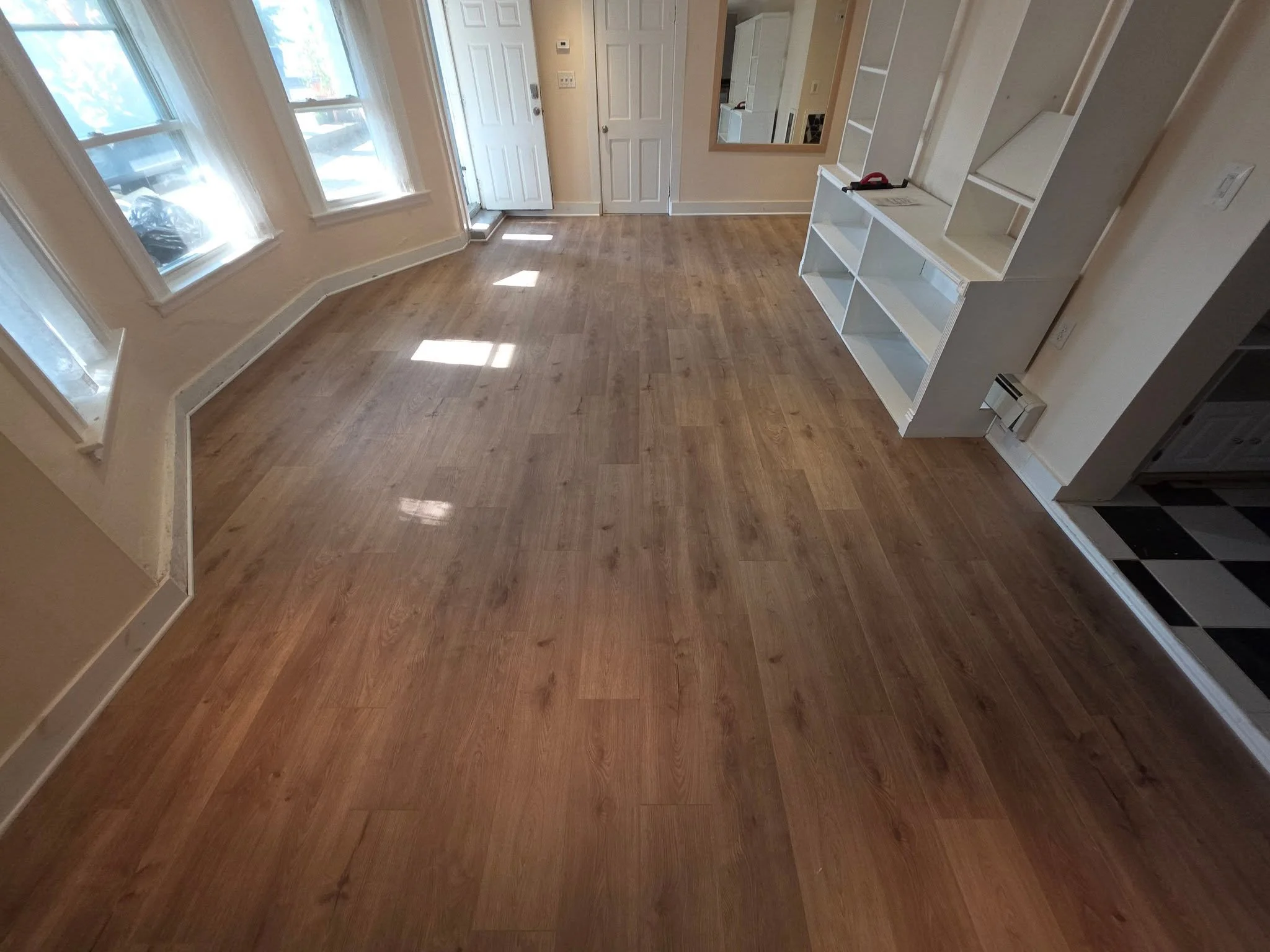 Empty room with hardwood floors, three windows letting in sunlight, a front door, and built-in white shelving units on the right side.