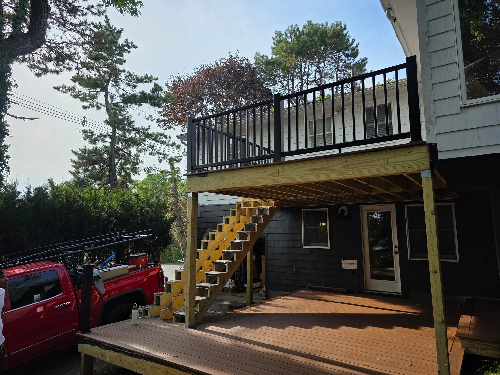 A two-story house with a balcony under construction. The second floor has a black metal railing, and a set of stairs leads up to the balcony. There's a wooden deck on the first floor, and a red pickup truck is parked nearby. The background features tall trees and power lines.