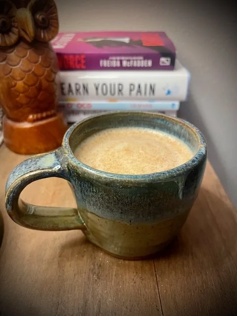 A ceramic mug filled with coffee on a wooden surface with books and a decorative owl figurine in the background.