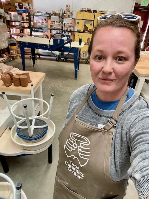 Woman taking a selfie in a pottery studio, wearing an apron, with pottery wheels and shelves of ceramic supplies in the background.