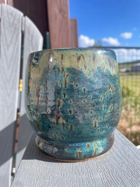 A ceramic pot with a glaze featuring shades of blue, green, and beige, placed outdoors on a white surface with a wooden fence, grass, and a blue sky with clouds in the background.