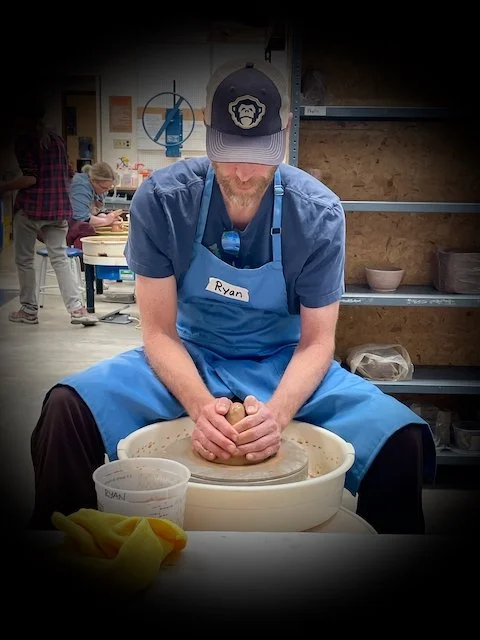 A man wearing a cap and apron throwing clay on a pottery wheel in a pottery studio.