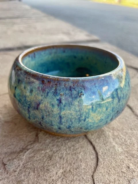 A small ceramic bowl with a blue and green glazed finish, sitting on a concrete sidewalk.