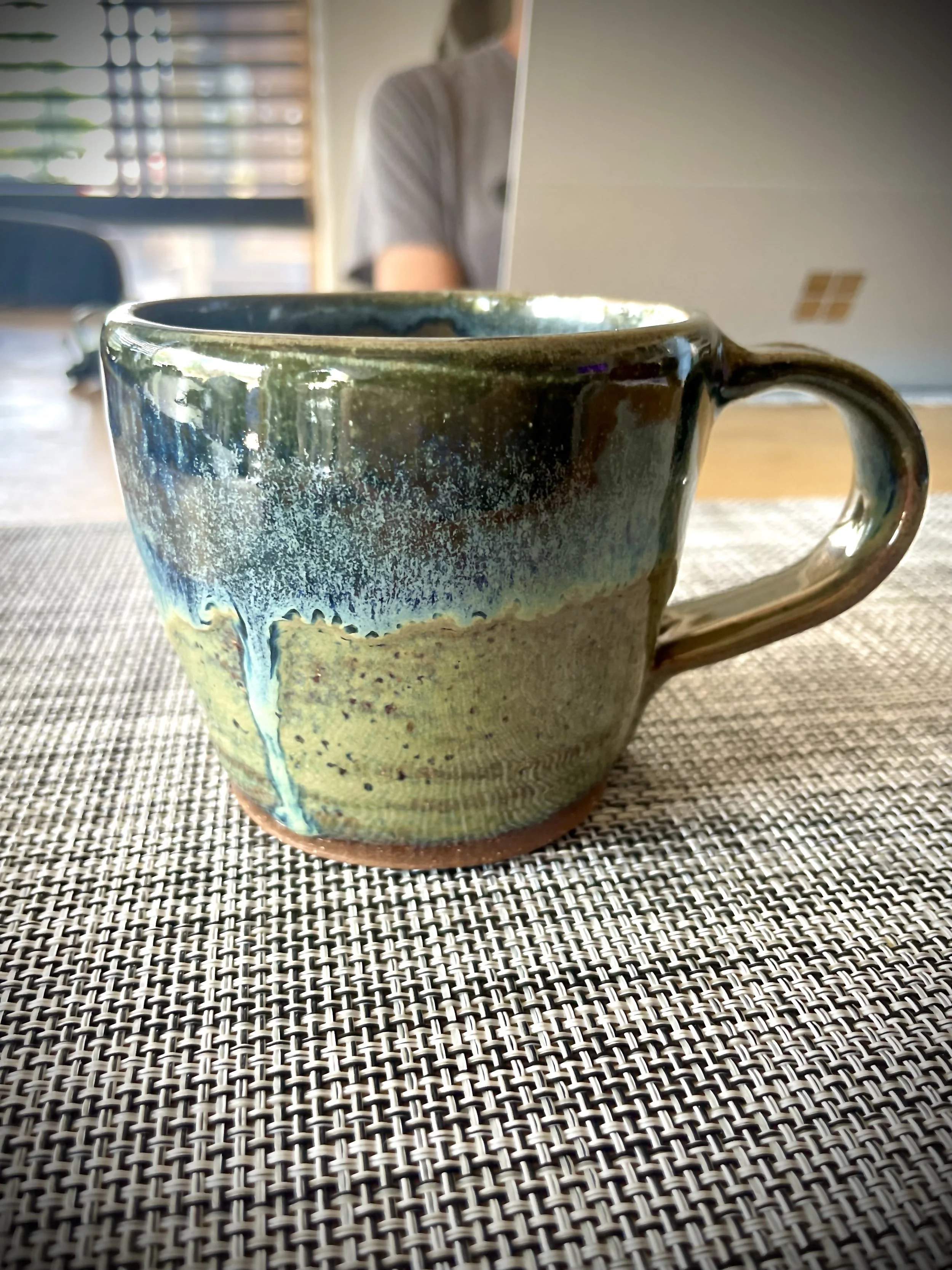 Close-up of a ceramic mug with a textured glaze in green and blue, placed on a woven placemat on a table.