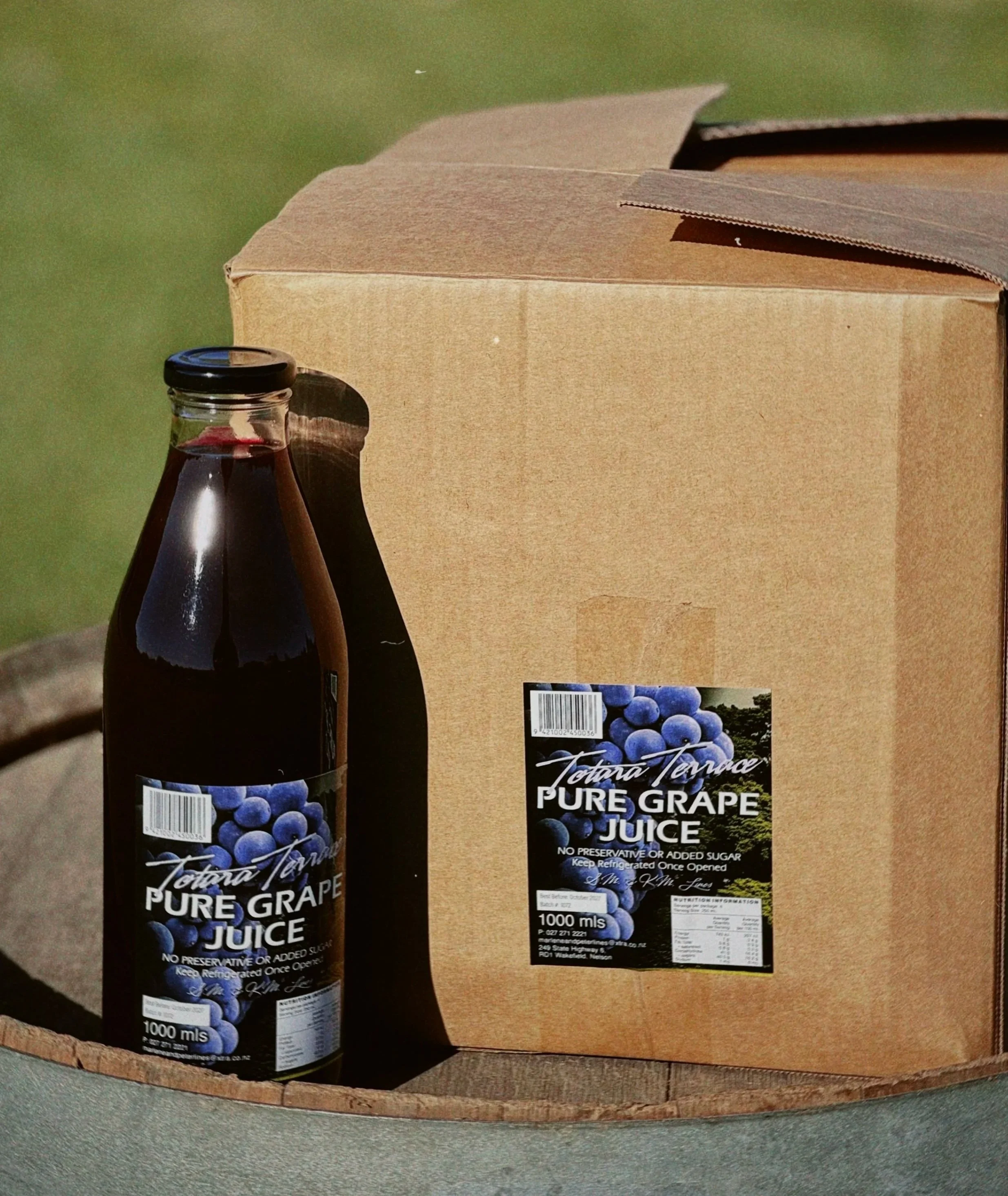 A cardboard box with a bottle of pure grape juice on top, both placed on a wooden surface. The bottle has a barcode, label with grapes, and nutritional information.