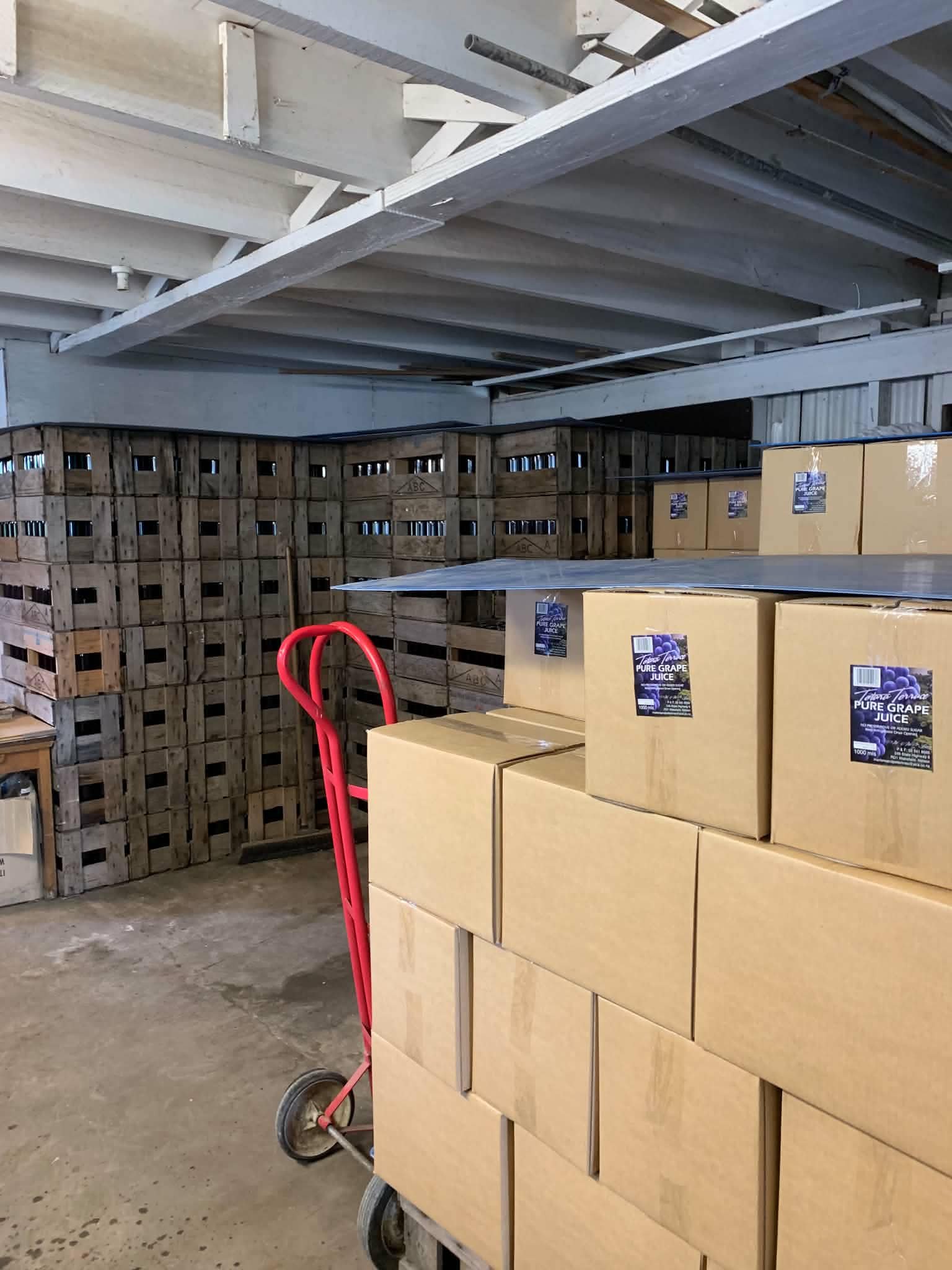 Rows of cardboard boxes labeled 'Pure Grape Juice' stacked on a flat surface inside a warehouse. Behind the boxes, there are wooden crates and a wooden shelving unit. A red hand truck is partially visible on the left side of the image.