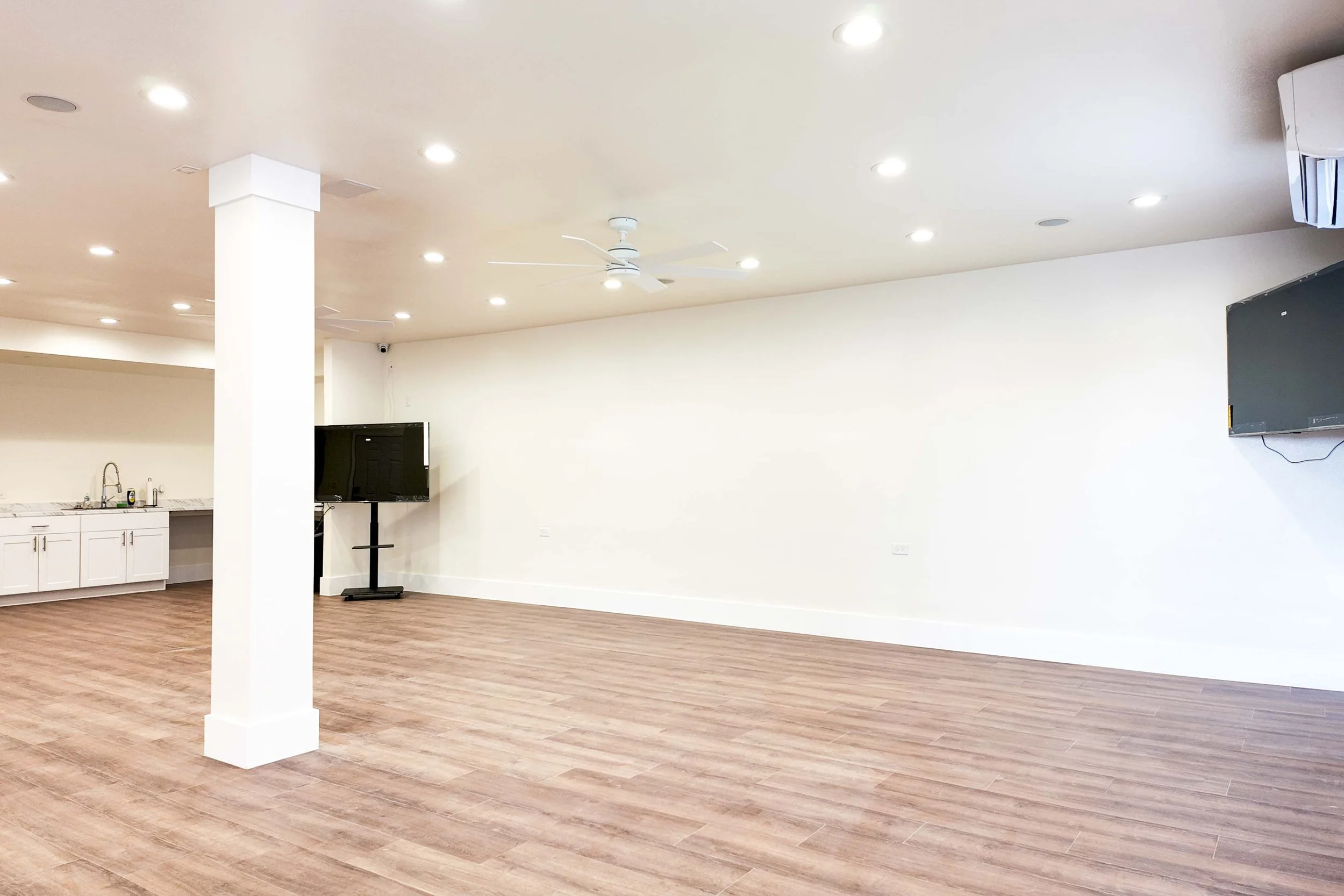 Empty spacious room with hardwood floors, white walls, ceiling fans, and two mounted TVs. Partially visible kitchen area with sink and cabinets.
