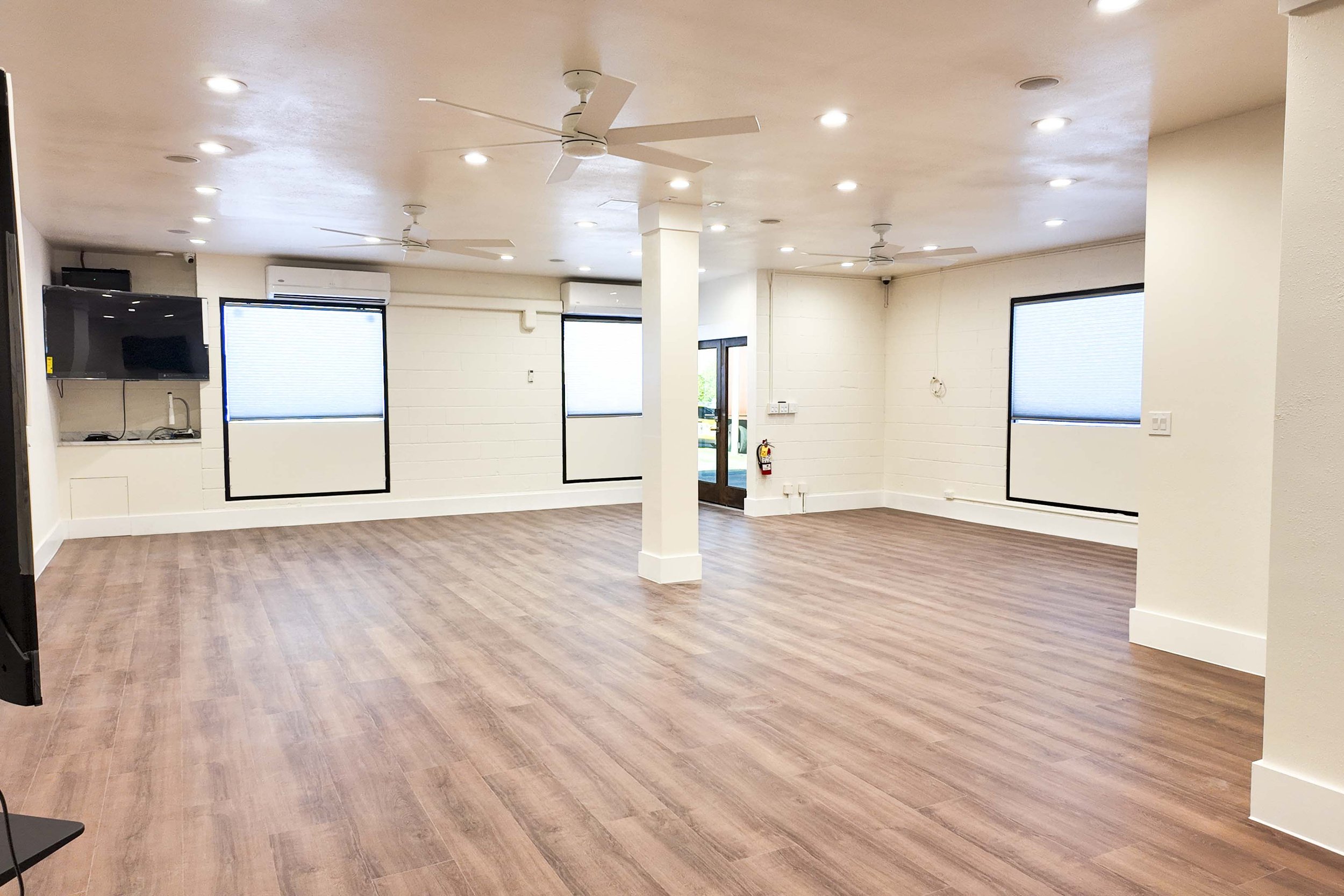 Empty room with wood-like flooring, white walls, four windows with blinds, ceiling fans, and recessed lighting.