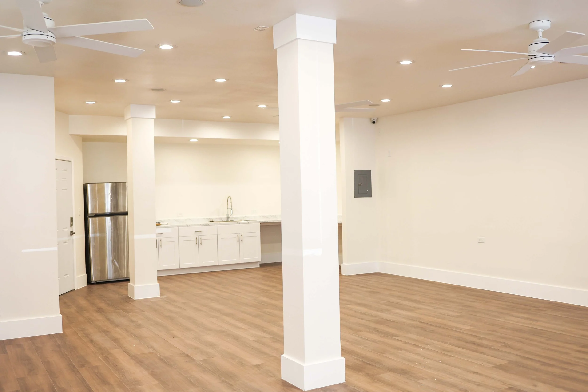 Interior view of an open-concept space with white walls, wood flooring, a small kitchen area with white cabinets, a stainless steel refrigerator, and ceiling fans.