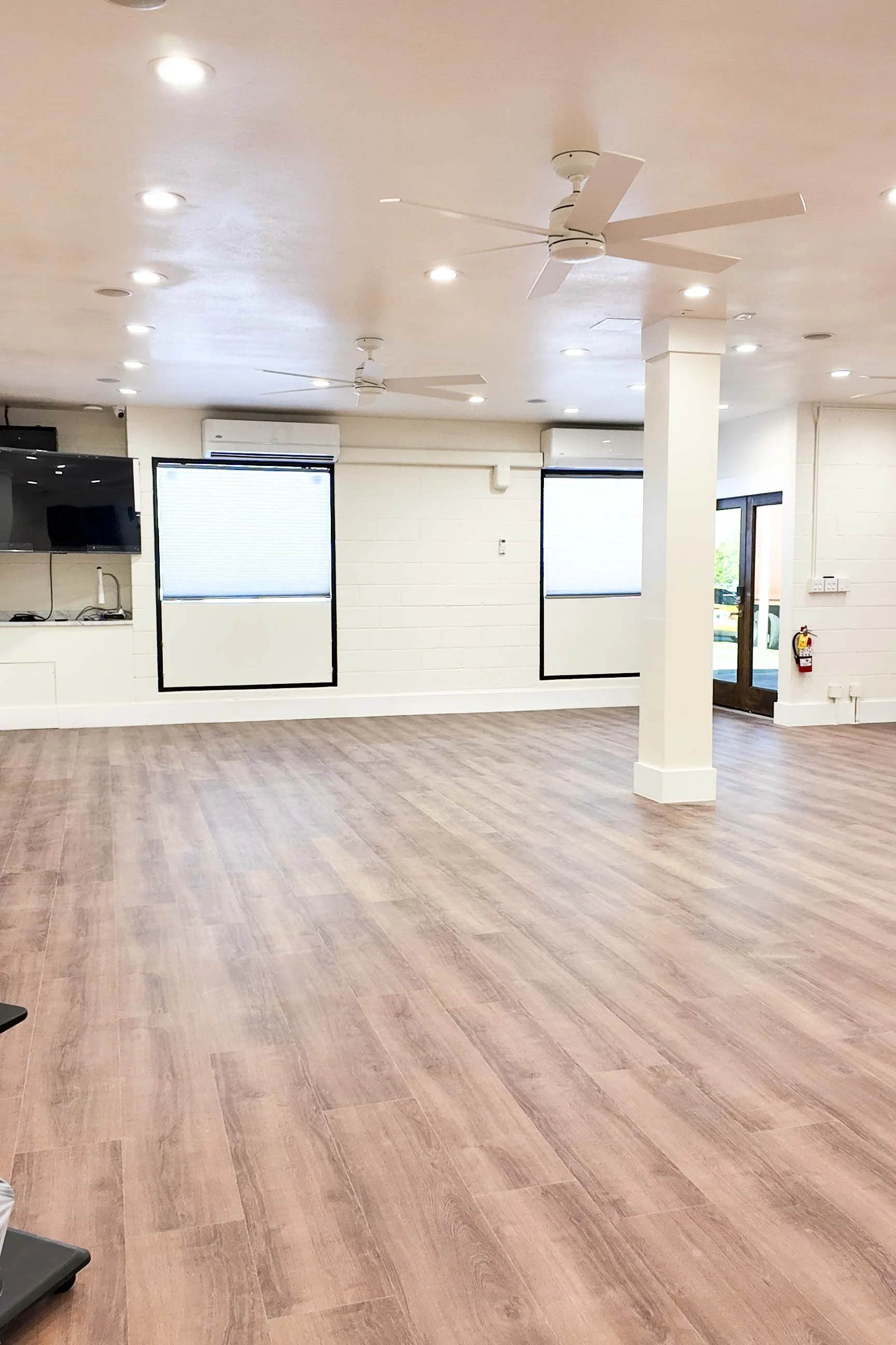 Empty room with wooden flooring, white walls, ceiling fans, and multiple windows with blinds.