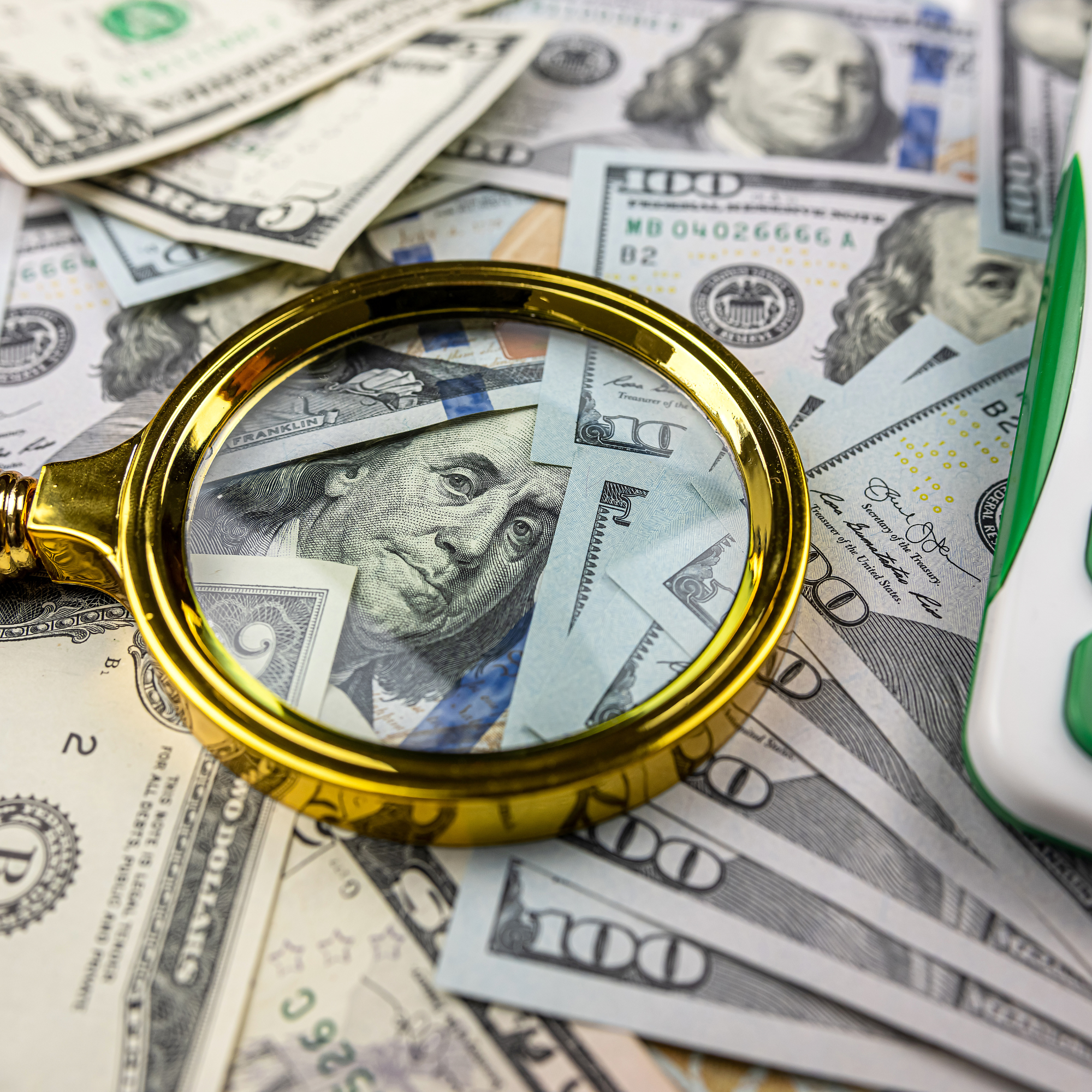 Various US dollar bills, including $20, $50, and $100, arranged with a gold-colored magnifying glass on top.