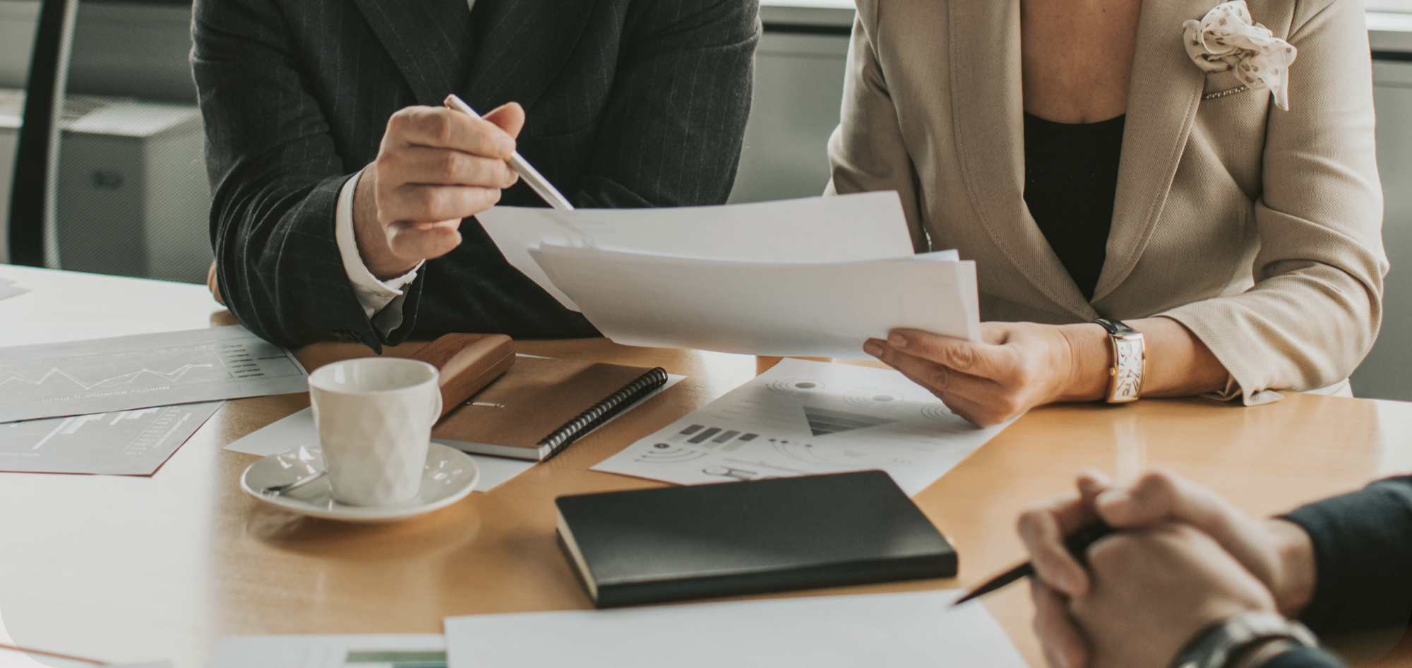 Two women in business attire reviewing documents at a conference table with charts and a coffee cup.